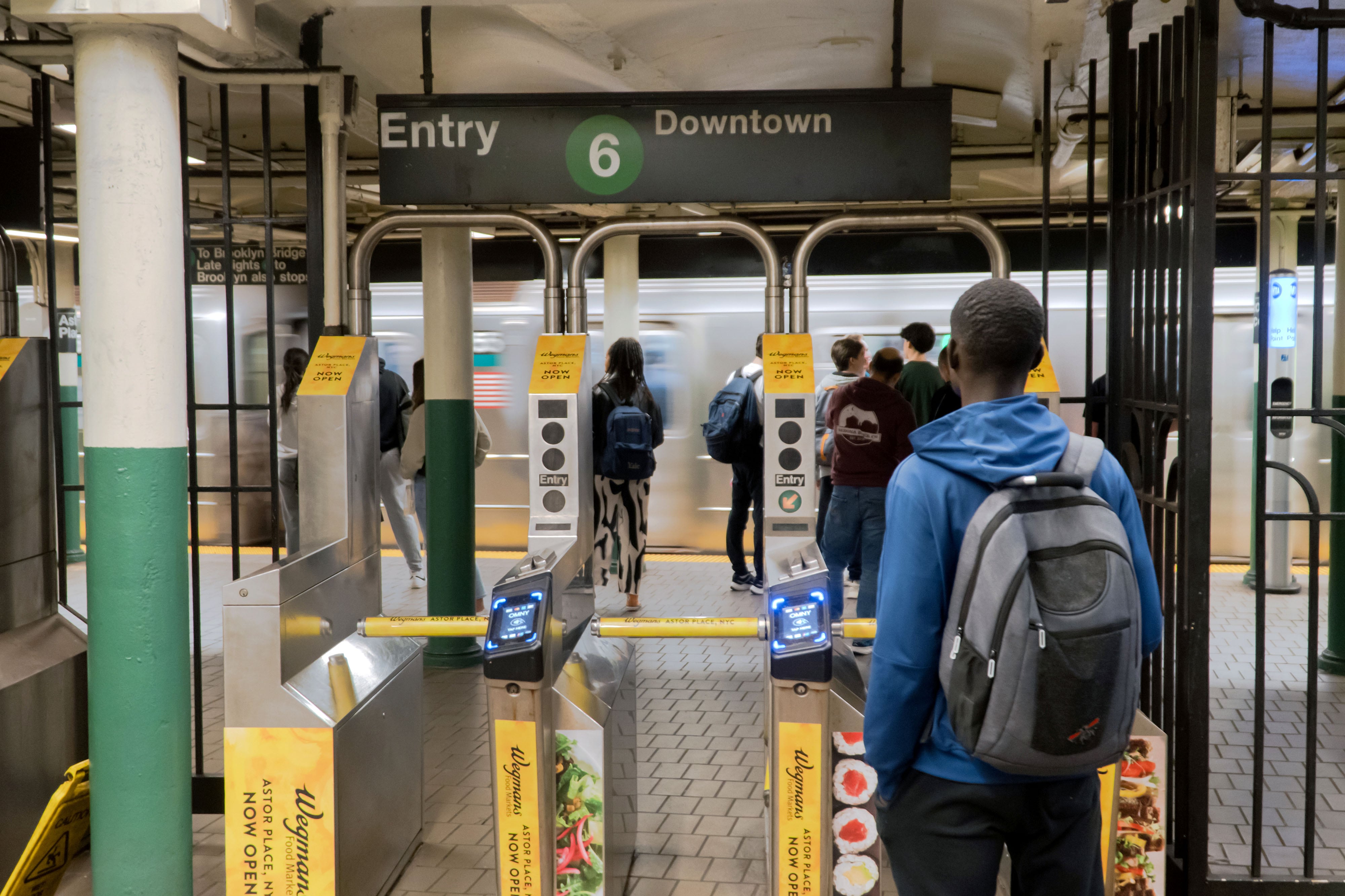 A young person wearing a blue hoodie and a dark backpack stands in front of the subway turnstiles with people standing in the background.