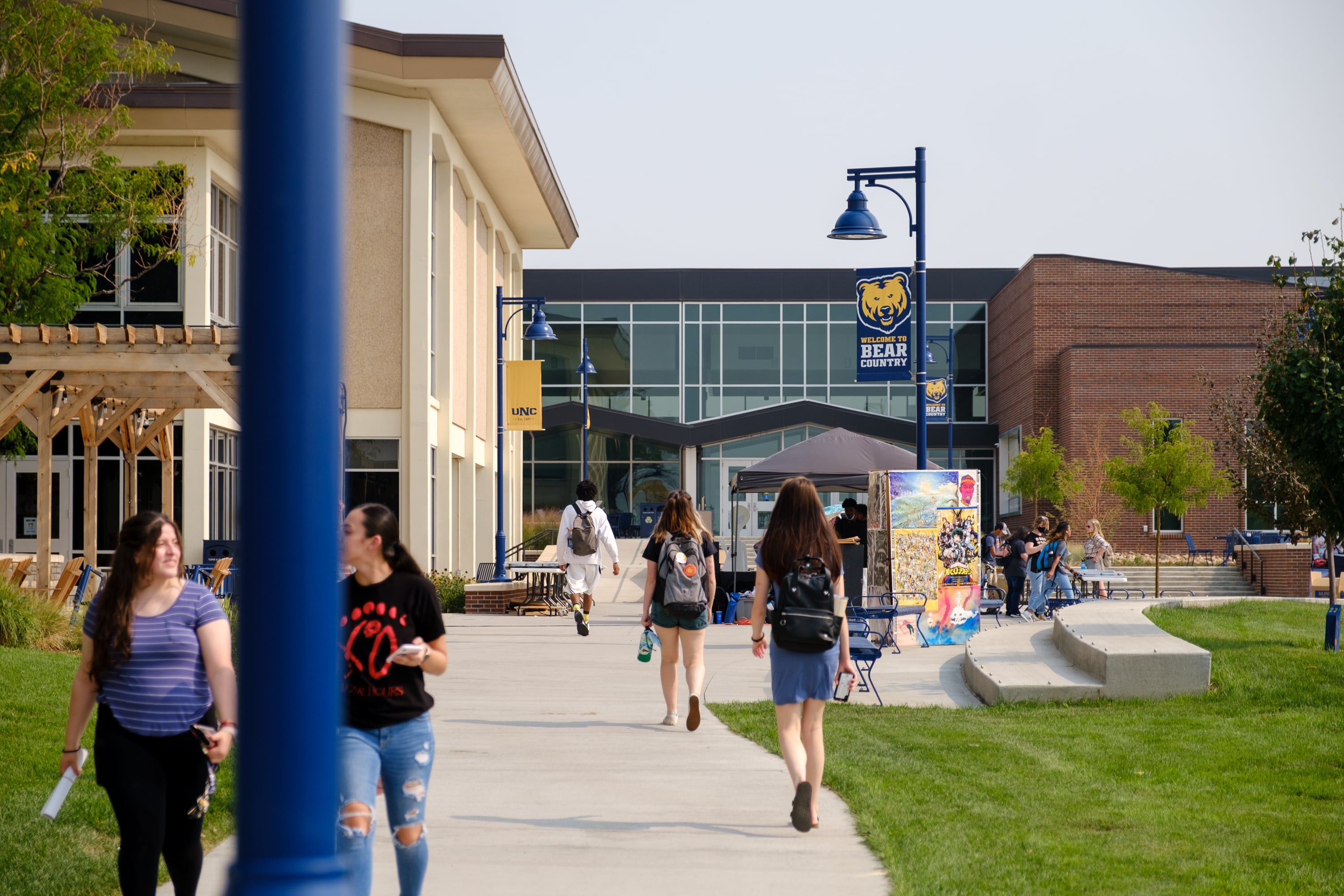 Students walk down a concrete sidewalk in between a grass lawn with a university building in the background.