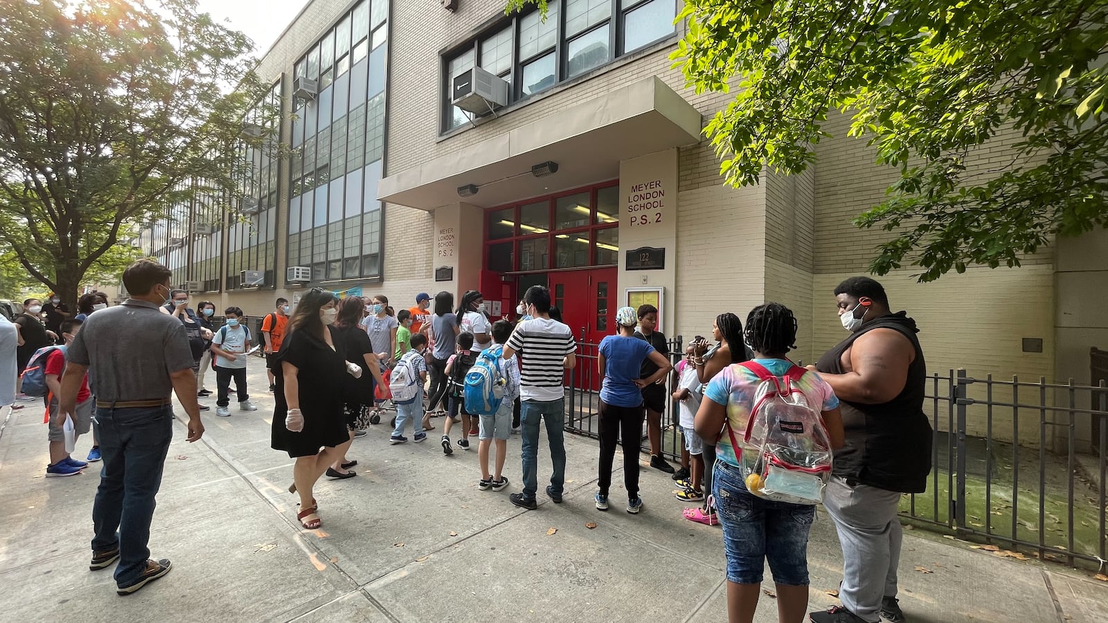 Several parents and their children wait outside of a school building before the start of a summer program. They are all wearing protective masks.