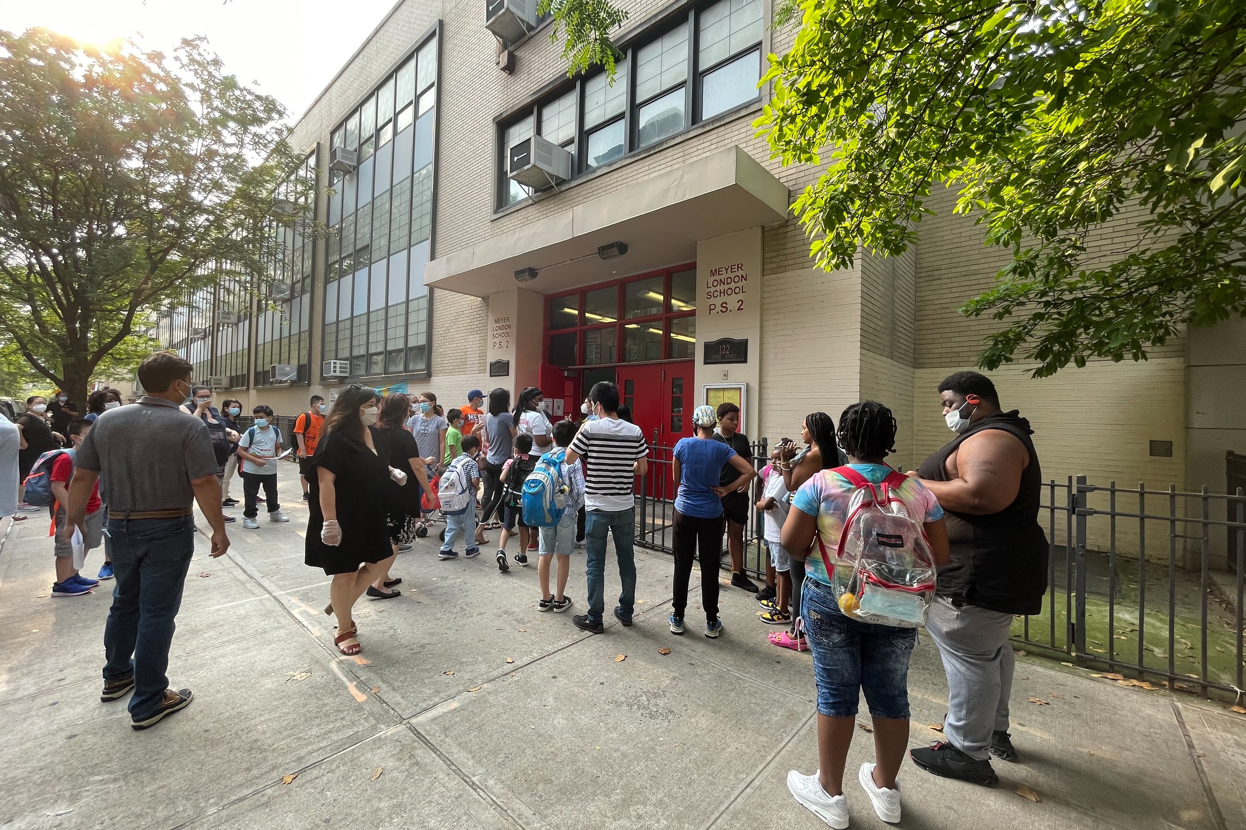 Several parents and their children wait outside of a school building before the start of a summer program. They are all wearing protective masks.