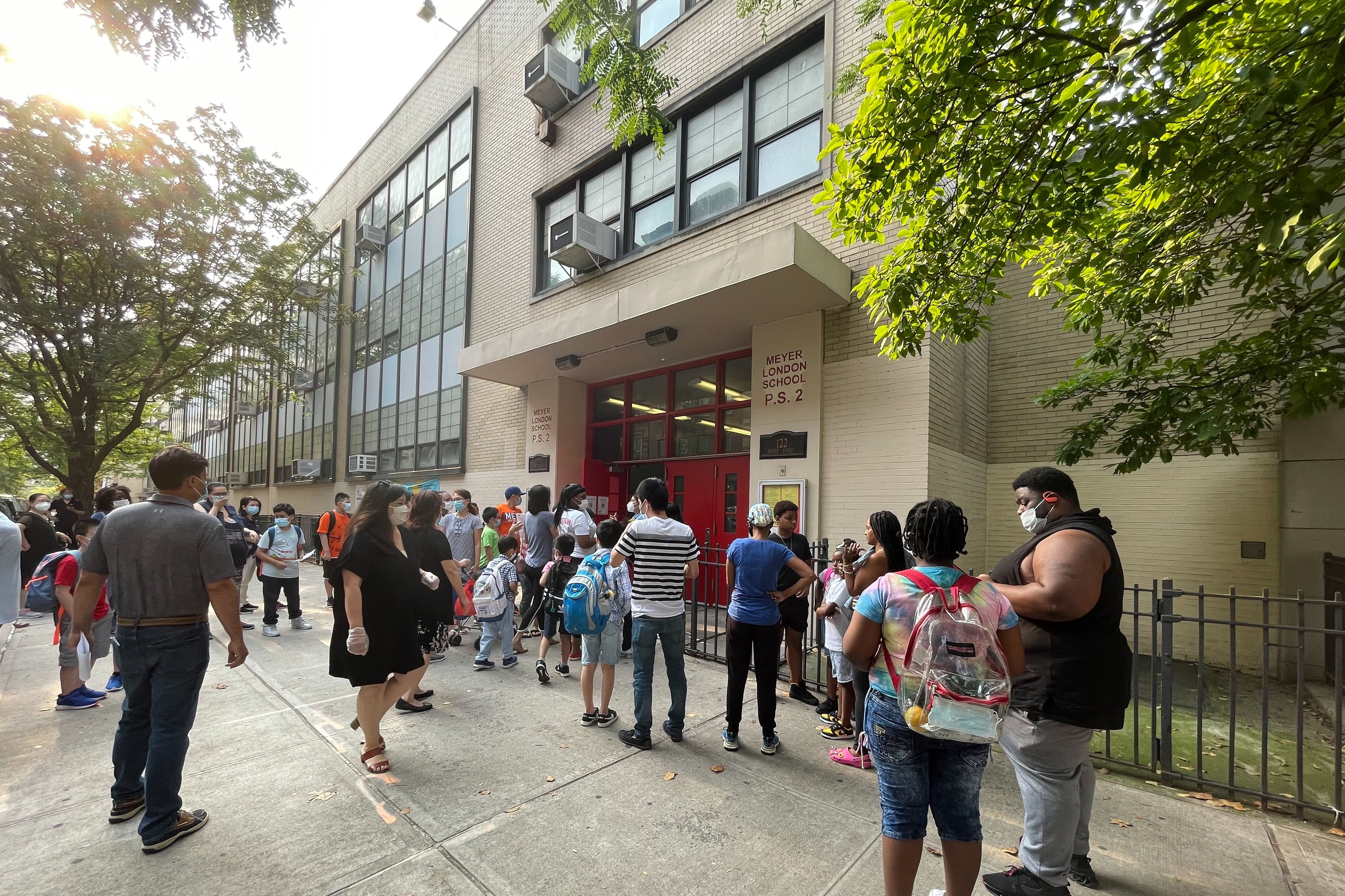 Several parents and their children wait outside of a school building before the start of a summer program. They are all wearing protective masks.