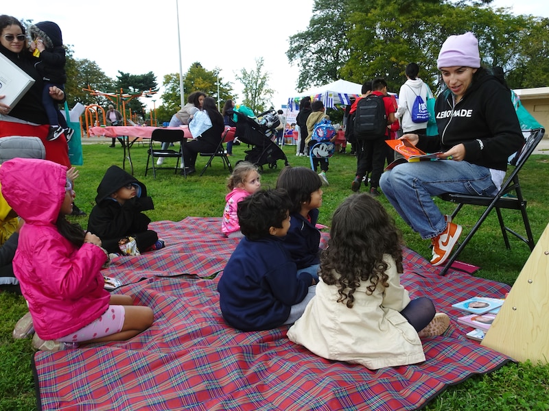 A person wearing a purple hat and a dark sweater sits on a chair reading a book to a group of young children all sitting on a red stripe blanket on the grass.