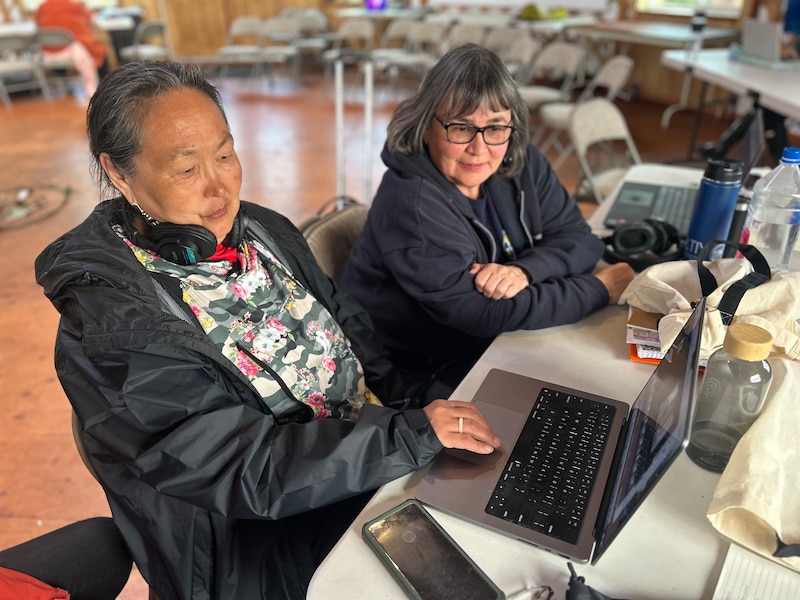 Two female teachers sit side by side with laptops open in front of them. One has a pair of headphones wrapped around her neck.