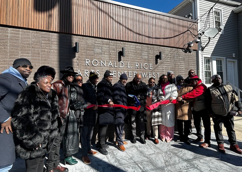 A line of more than a dozen adults dressed in cold weather clothing cut a red ribbon outside of a brown brick building.