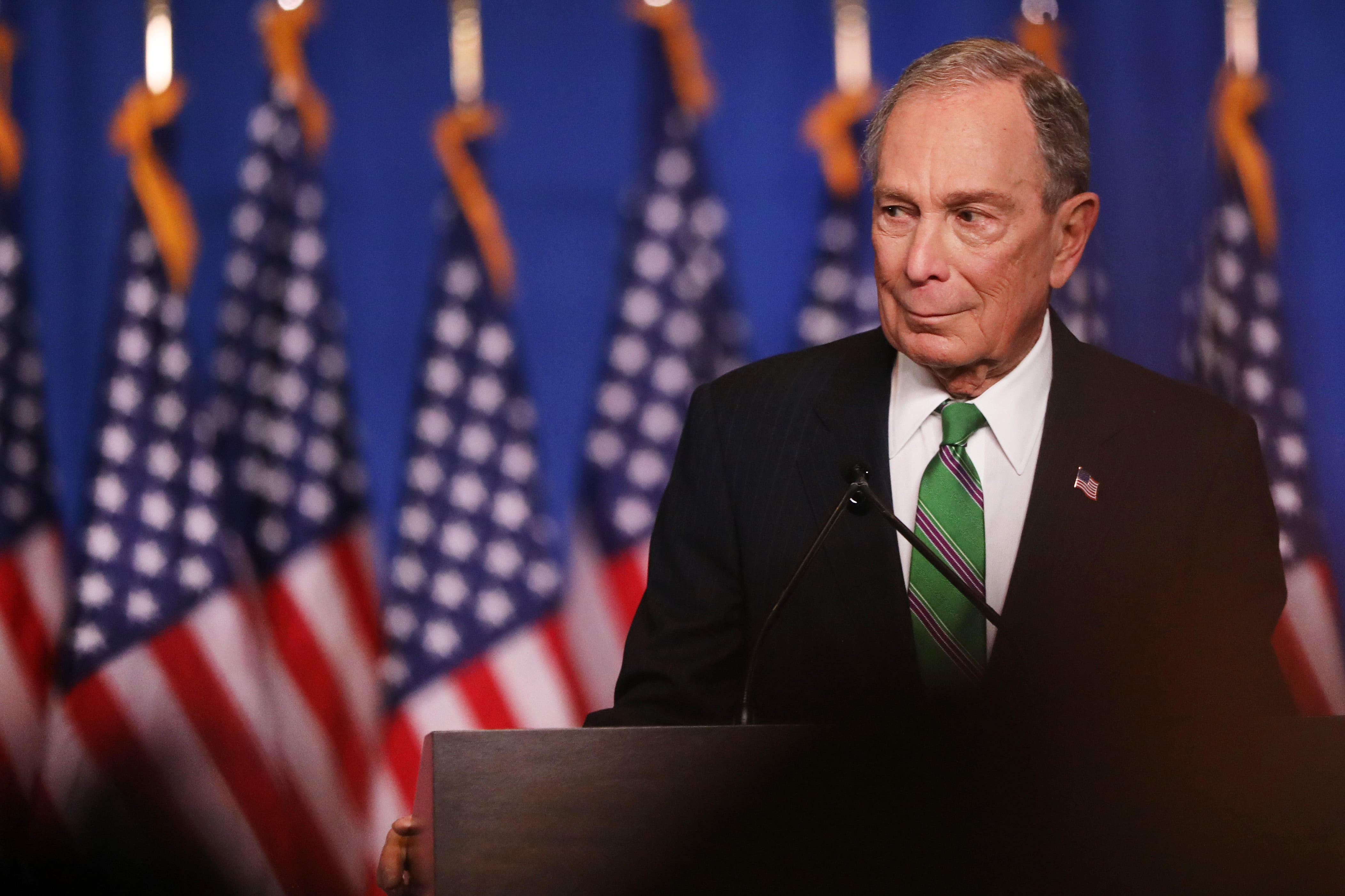 Michael Bloomberg, wearing a green tie, stands at a podium in front of a row of American flags.