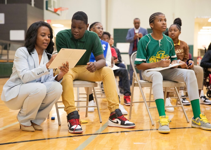 An adult in a light blue suit kneels down next to a student sitting in a chair.