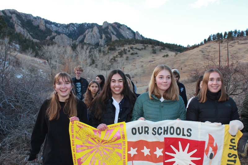 A photograph of four women standing side by side holding a sign with a mountain range in the background.