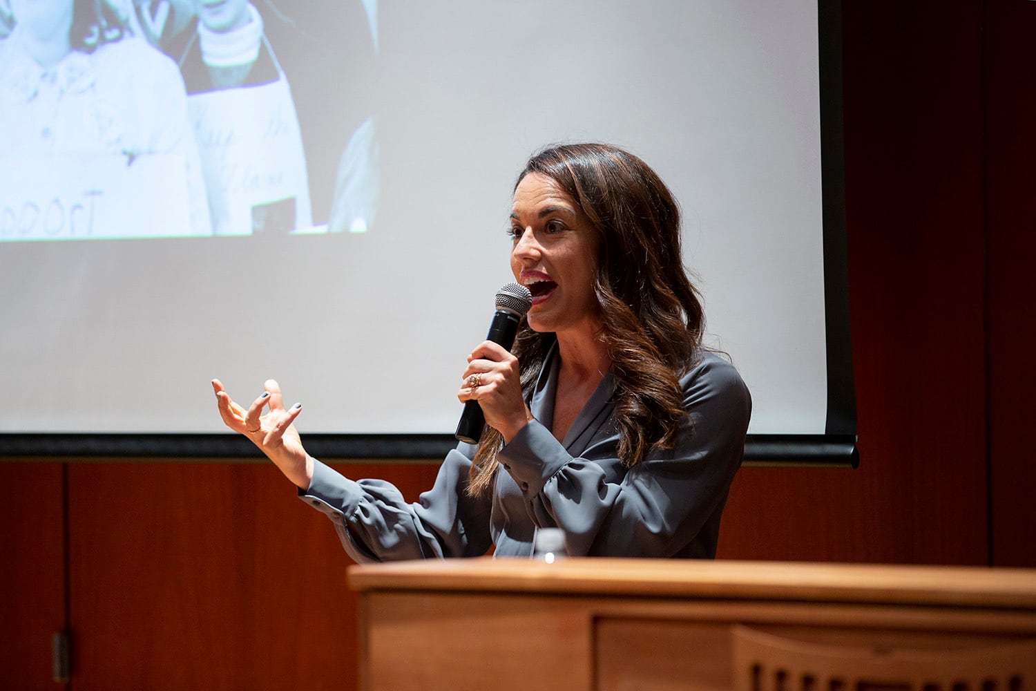 A white woman with dark hair stands in front of a wooden podium.
