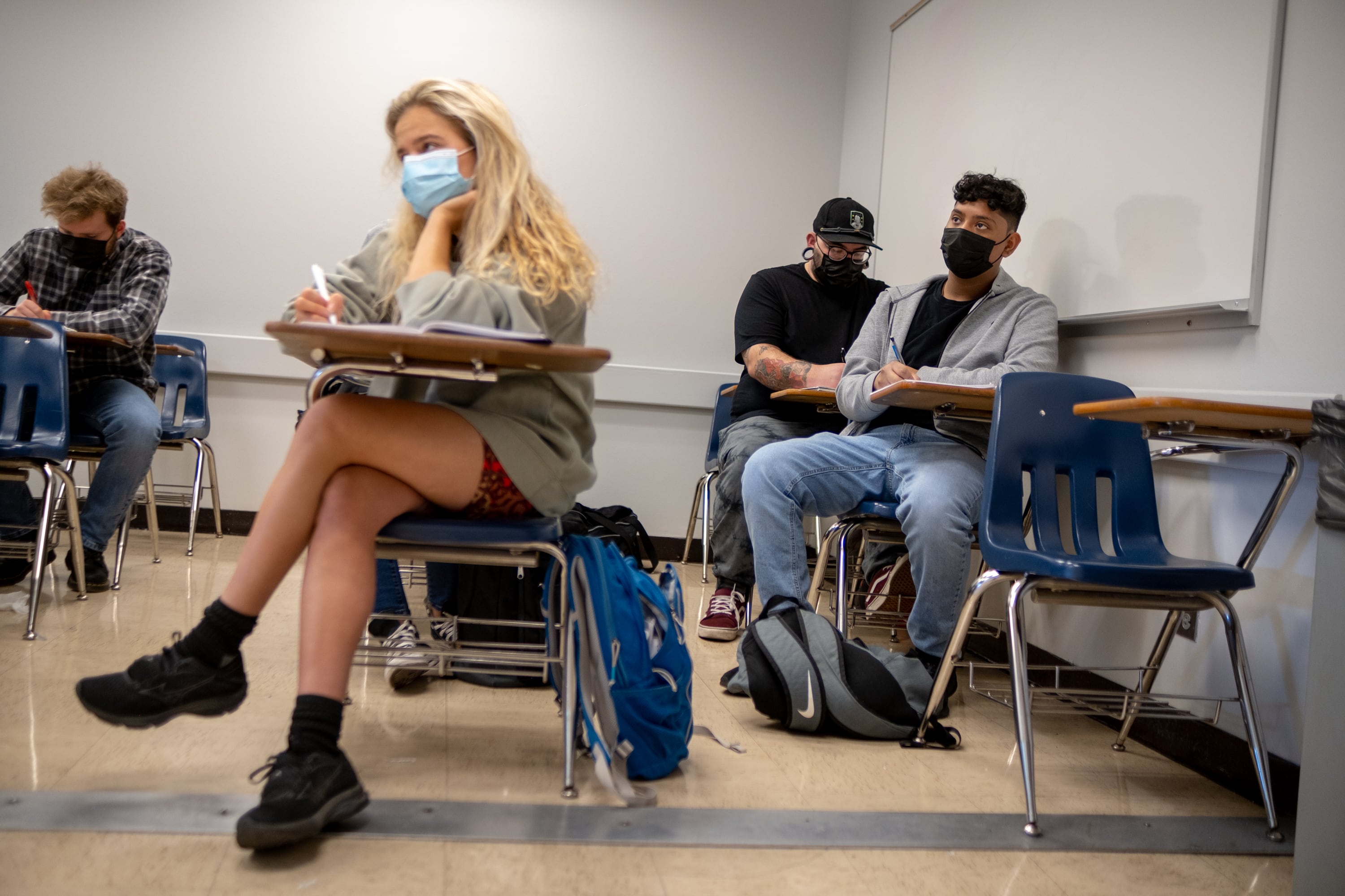 A student with long blonde hair and black shoes sits at a desk in the foreground of a classroom with white boards, while three other students wearing masks sit at desks in the background.