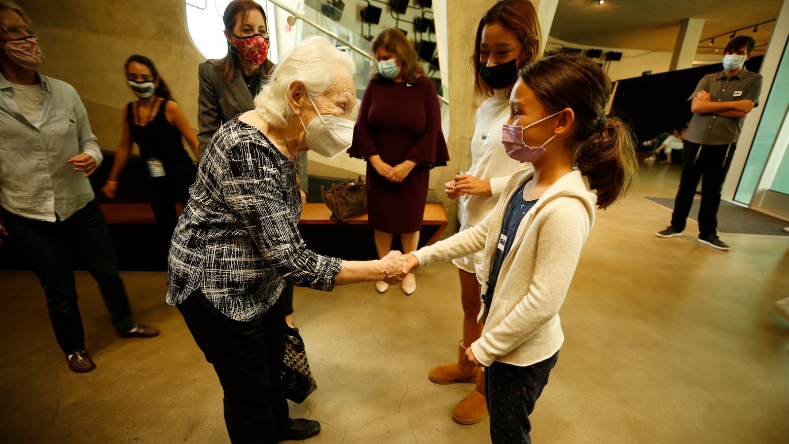 An elderly woman shakes hands with a girl and they make eye contact both wearing masks