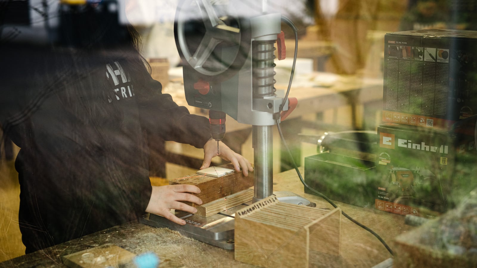A view through a window shows a student's pair of hands working on a drill.