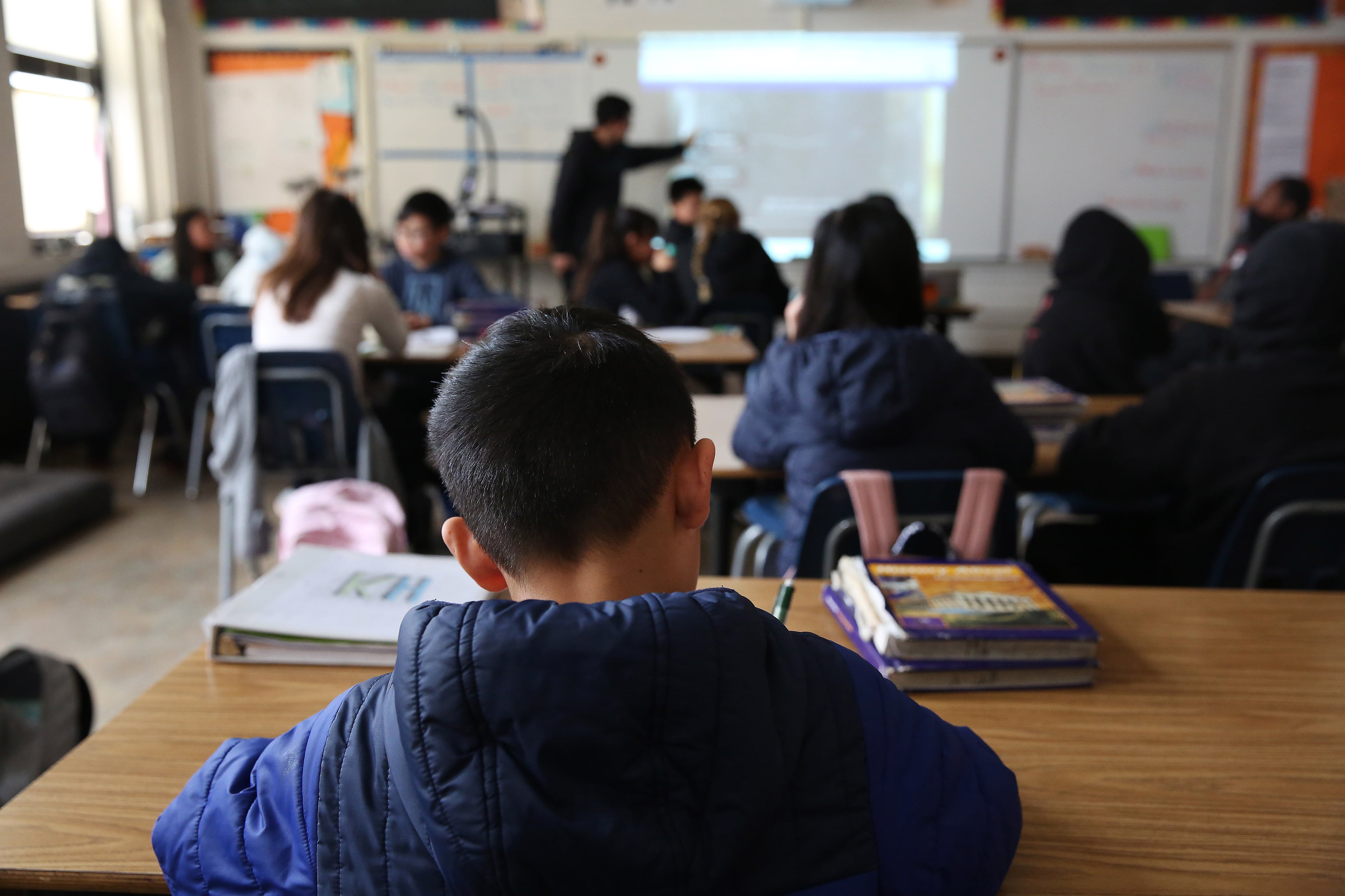 A view of the back of a middle schooler's back while they take notes with a classroom and a teacher in the background.