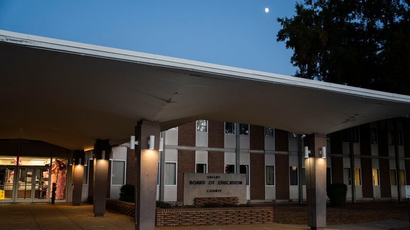 The front entrance to a red brick building with a large white roof at night with trees and the moon in the background.