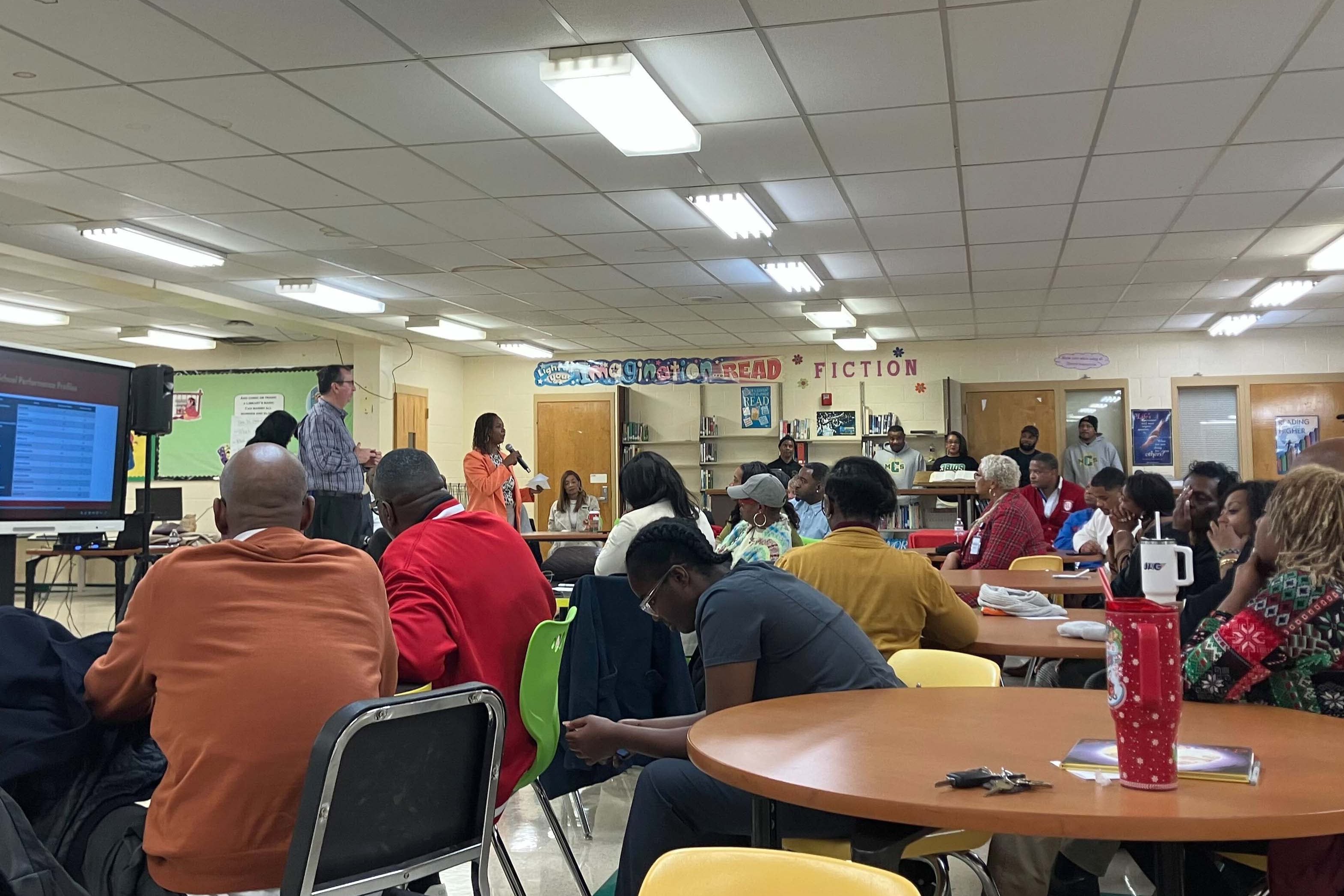 A photograph of a school cafeteria in a middle school full of community members sitting at tables listening to a person speak.
