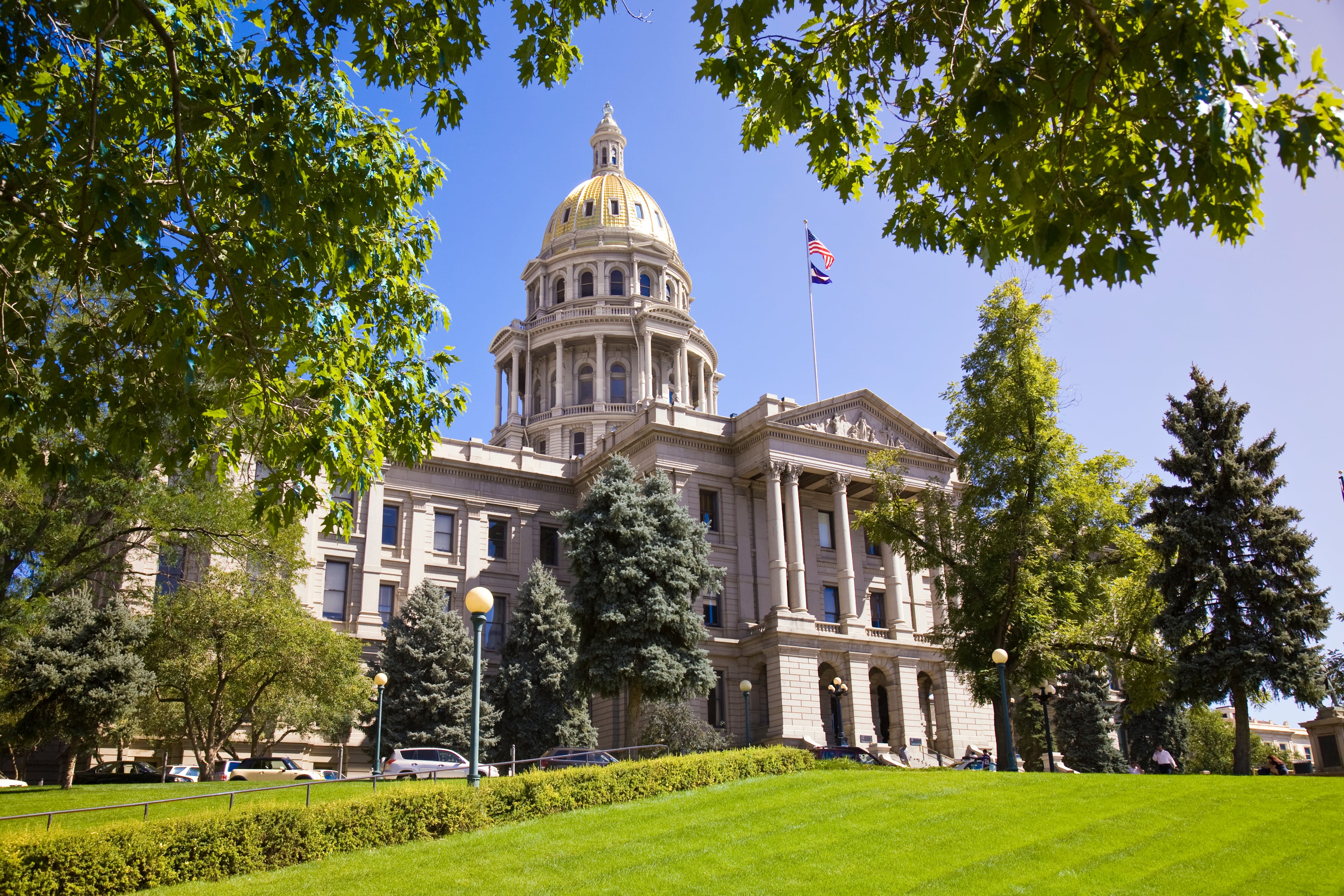 The Colorado State Capitol stands behind lush green trees on a sunny day. The flags of the United States and Colorado hang above large columns, with it's golden dome in the center of the frame.
