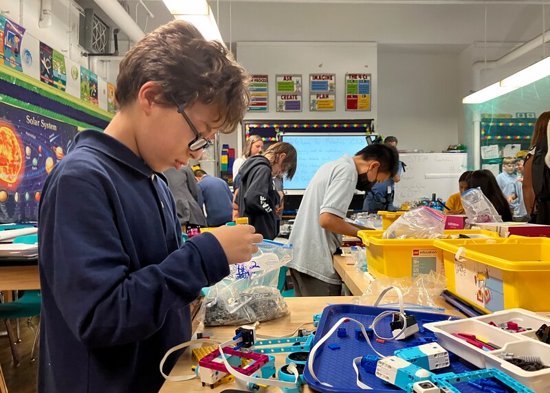 A young student wearing glasses works on a project during class with students working around tables in the background.