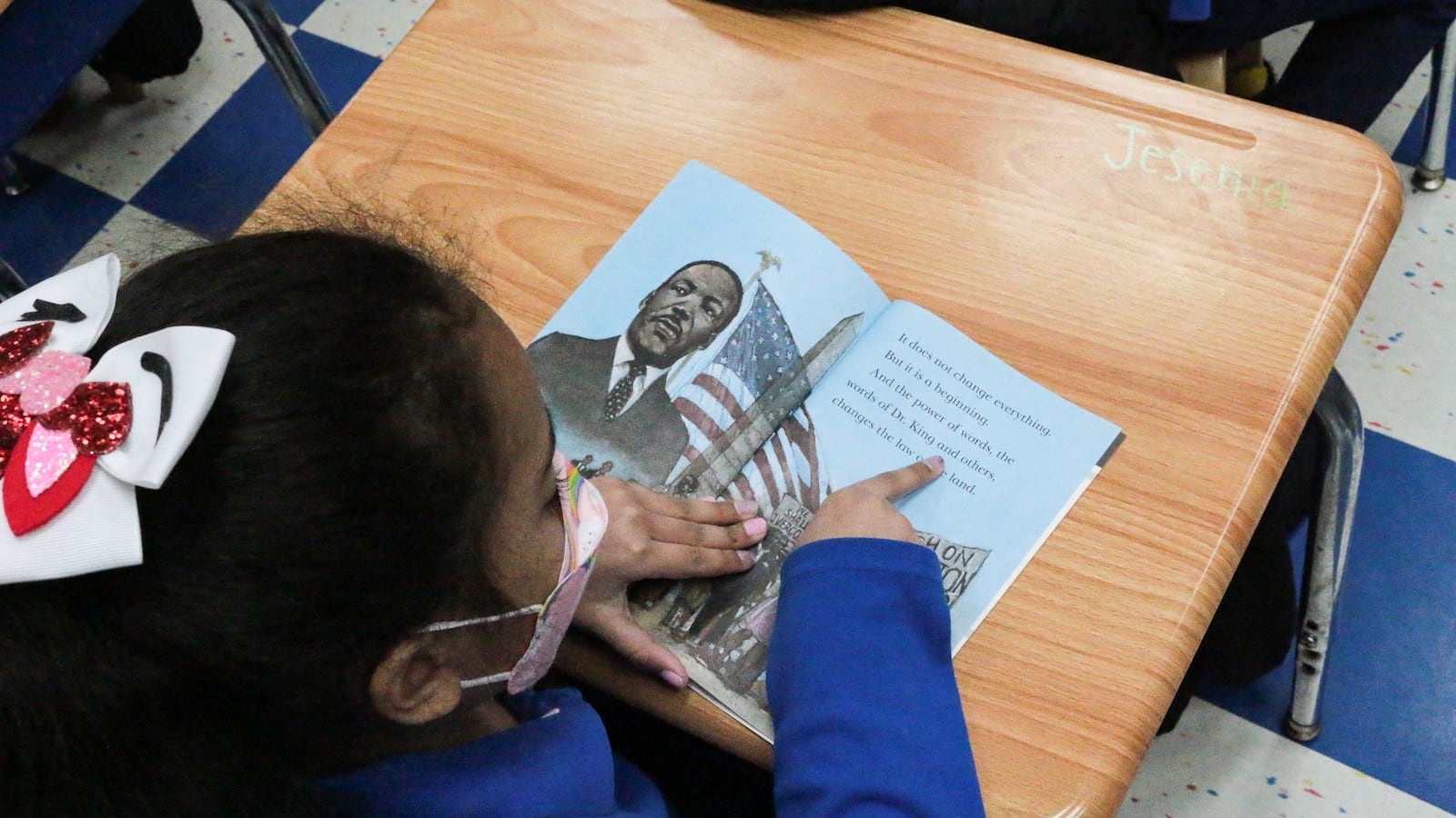 A girl leans over a book, pointing at a word with her finger.