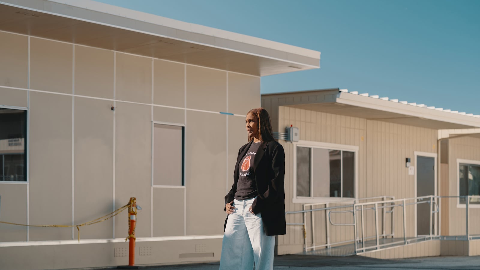 A photograph of a Black woman standing for a portrait outside of a school building on a sunny day.