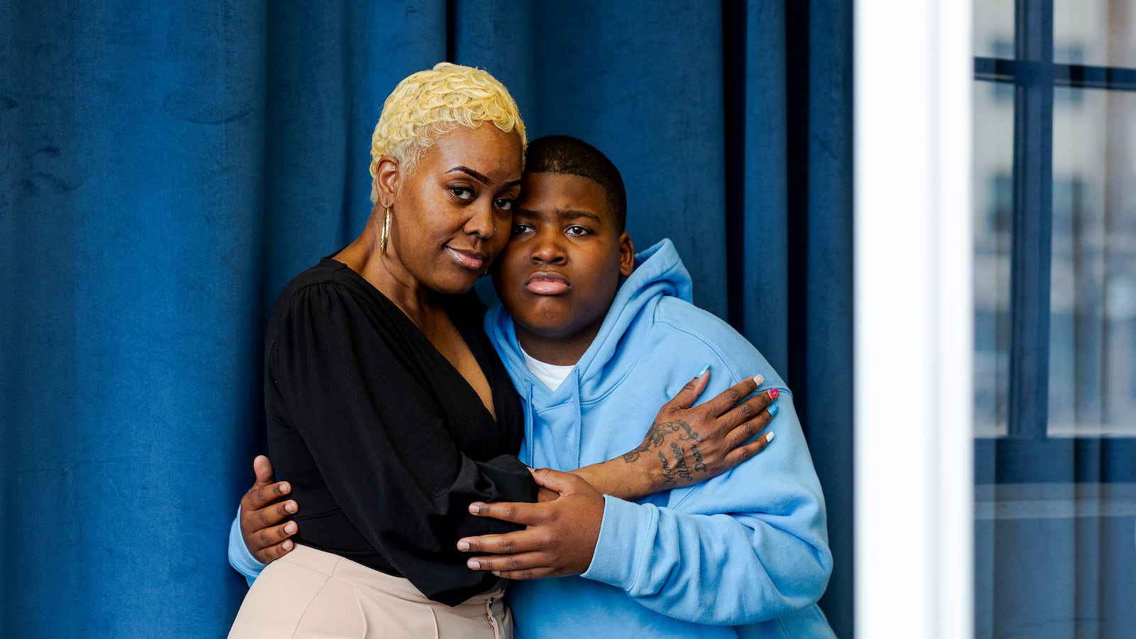 A Black mother and son pose for a portrait standing next to each other embracing against a blue curtain.