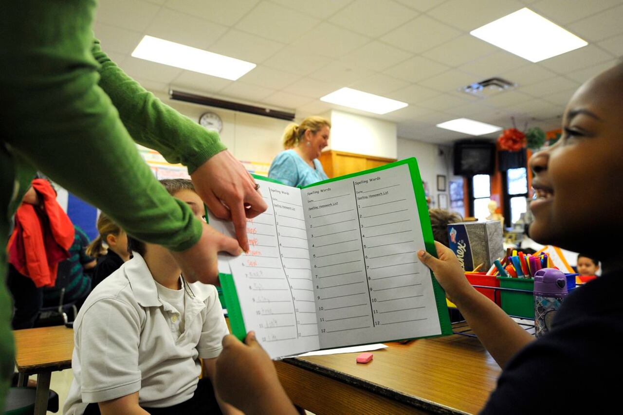 A photograph of a young Black student holding a worksheet folder up to an adult teacher who is pointing to part of the paper. The student is smiling and sitting at their desk in a classroom.