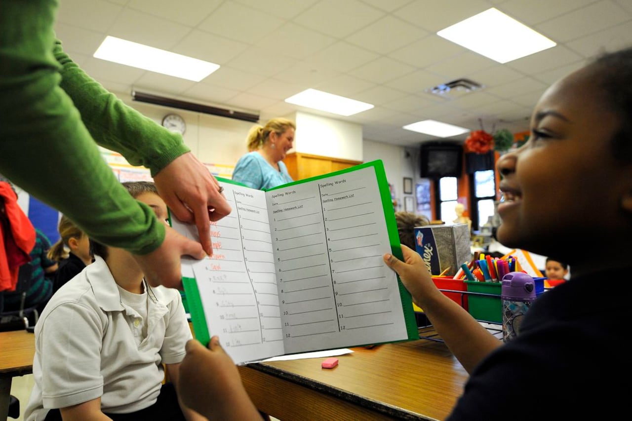 A photograph of a young Black student holding a worksheet folder up to an adult teacher who is pointing to part of the paper. The student is smiling and sitting at their desk in a classroom.