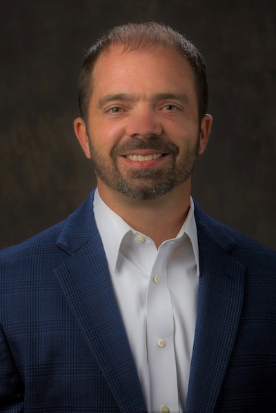Headshot of a man with a beard. He wears a white shirt and a dark blazer.