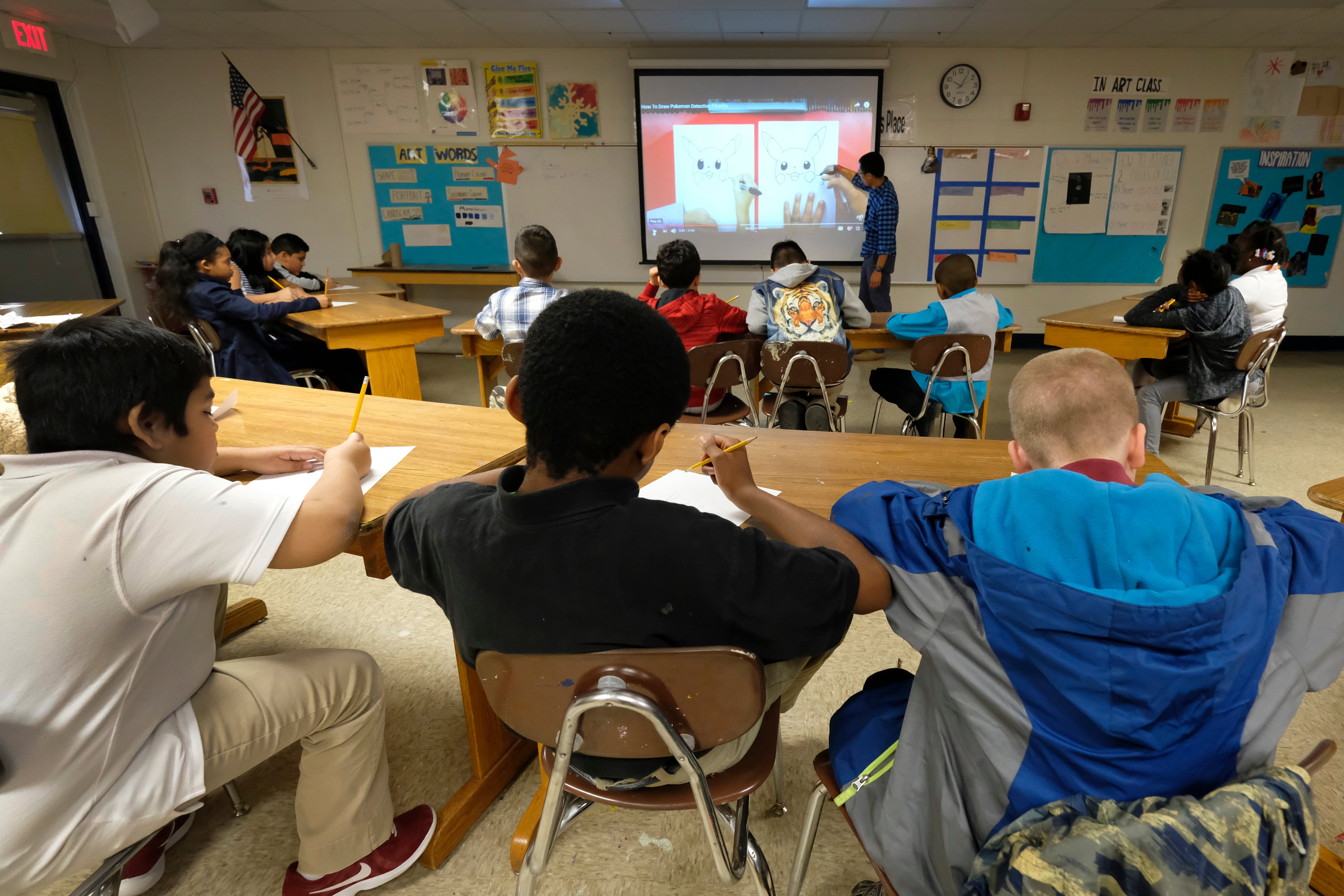 A row of three students sit at the back of the class, facing a projector screen where a teacher is showing a lesson.