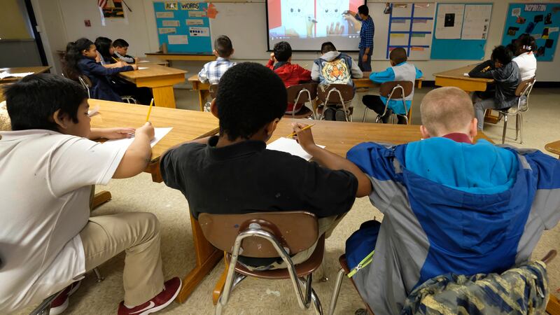 A row of three students sit at the back of the class, facing a projector screen where a teacher is showing a lesson.