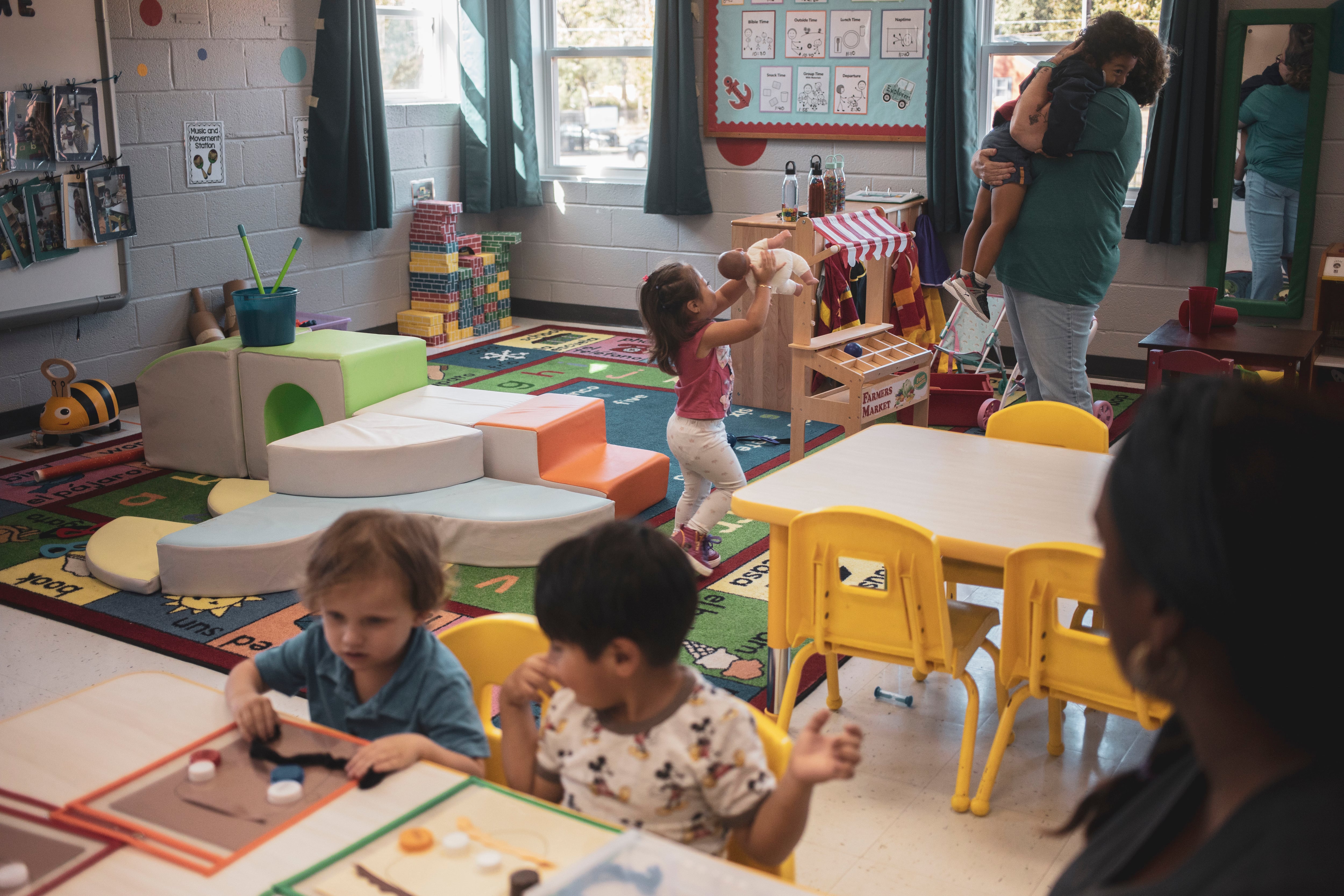 A teacher holds a child in the background of the classroom as a little girl runs up to her with a doll. Two students play together at a table in the foreground with another teacher.
