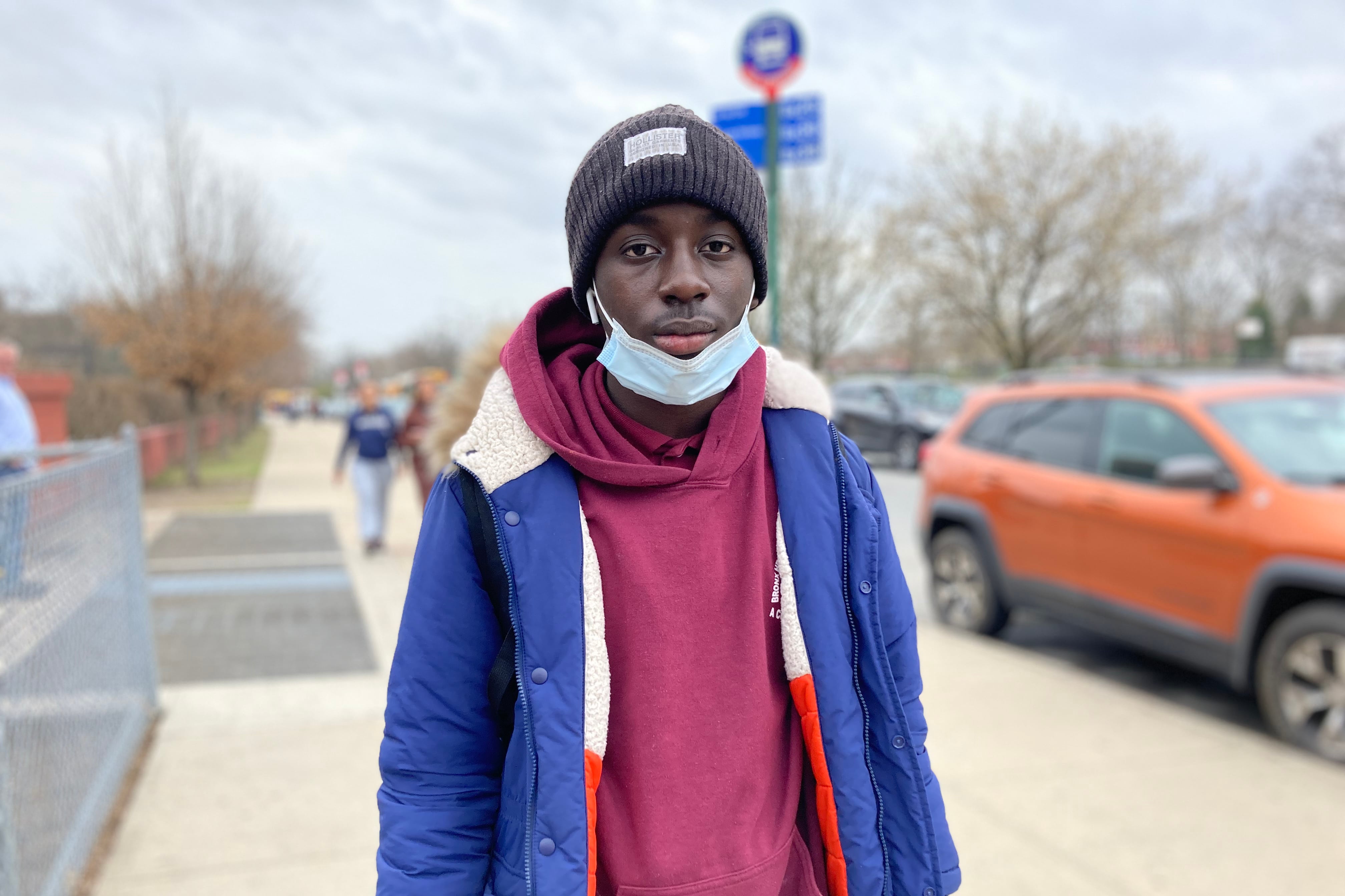 A high school students stands on a sidewalk near a bus stop.