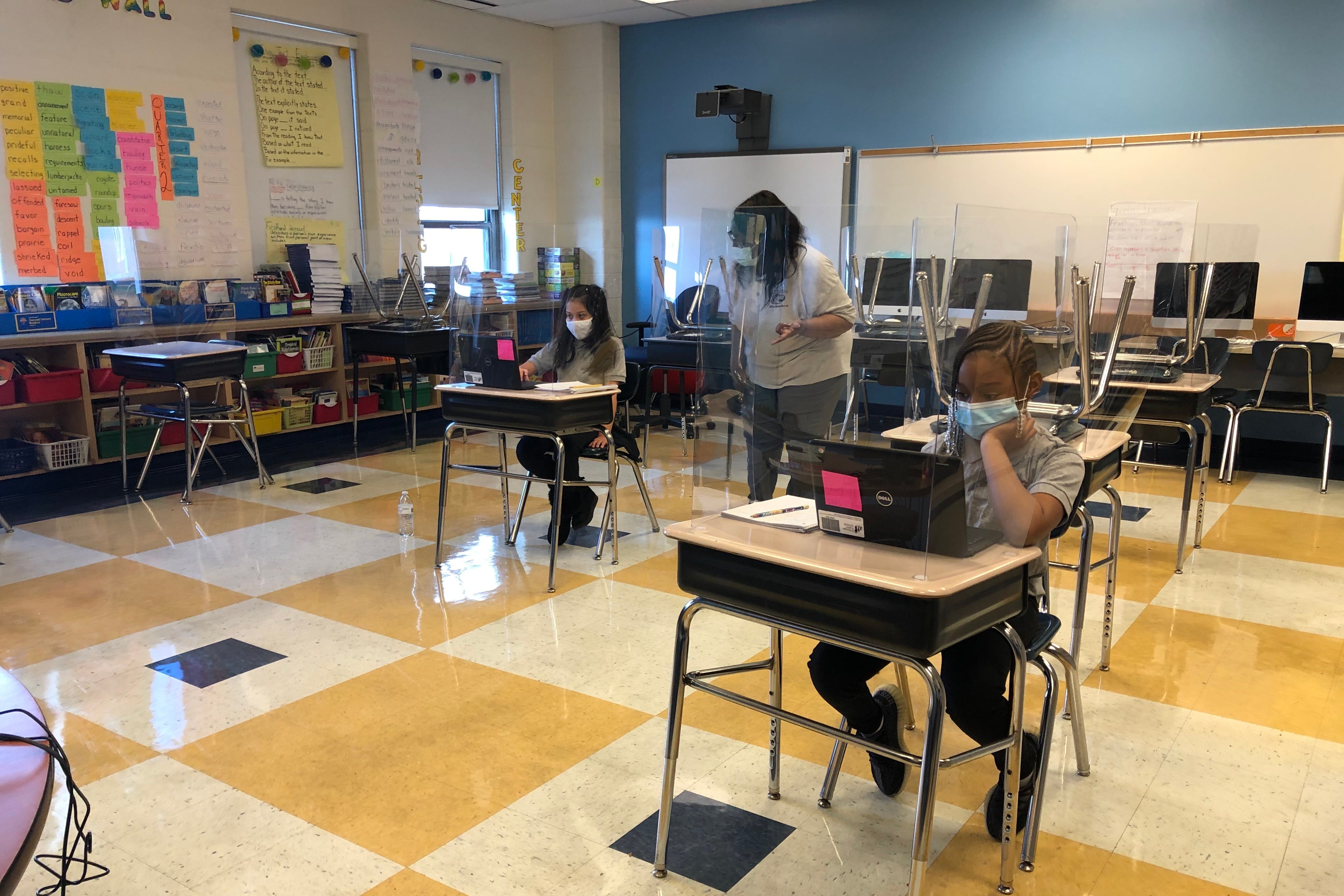 Students wearing face masks sit at desks behind plastic shields in a Chicago classroom with a yellow, cream, and black checkerboard floor and a blue wall. They are working on laptop computers as a teacher wearing a white top watches.