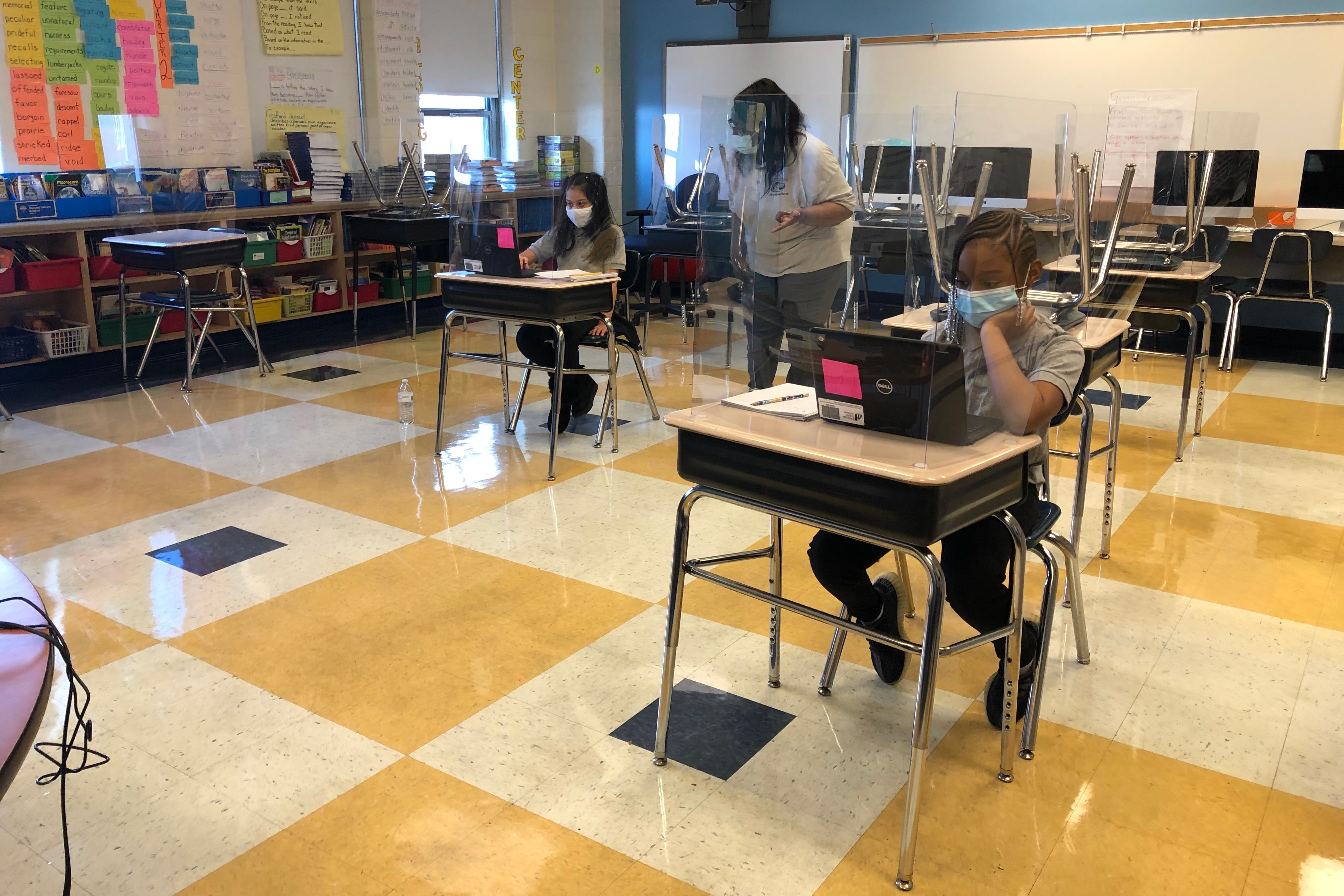 Students wearing face masks sit at desks behind plastic shields in a Chicago classroom with a yellow, cream, and black checkerboard floor and a blue wall. They are working on laptop computers as a teacher wearing a white top watches.