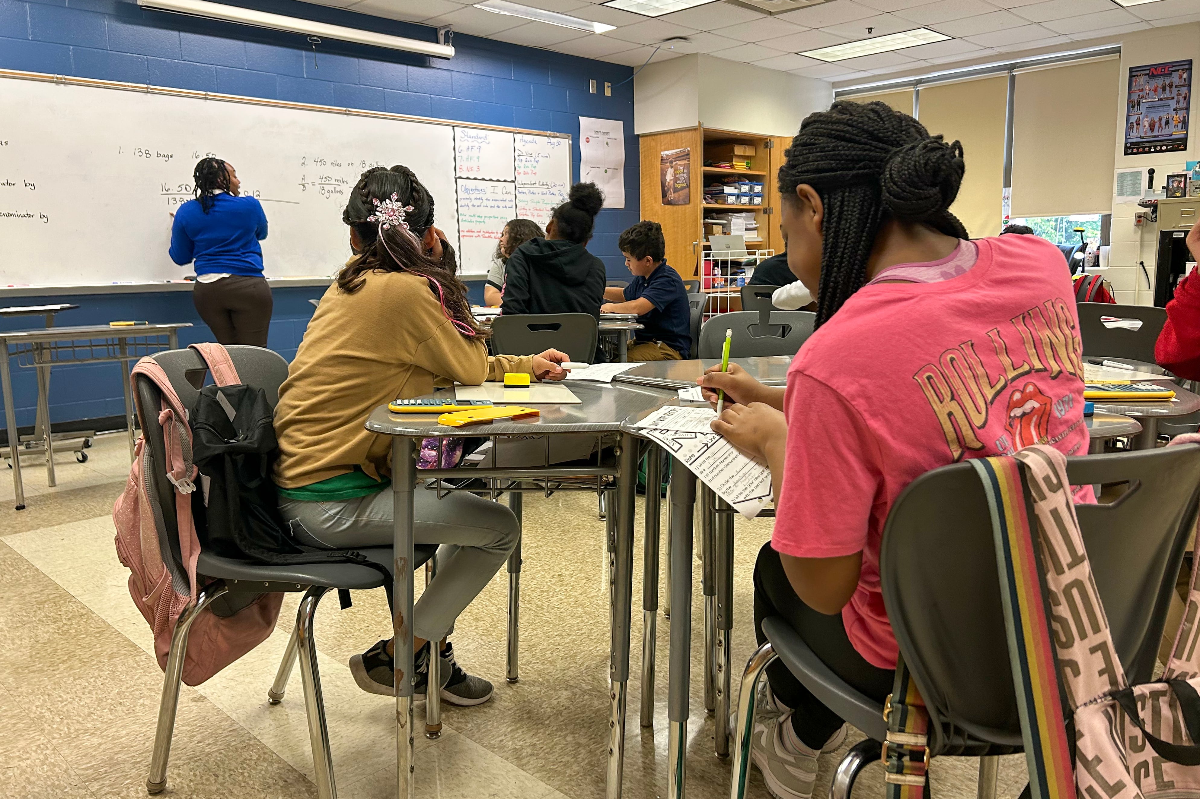 A student wearing a pink shirt sits at a table with other students in a classroom. In the background, a teacher in a blue top writes on the whiteboard.