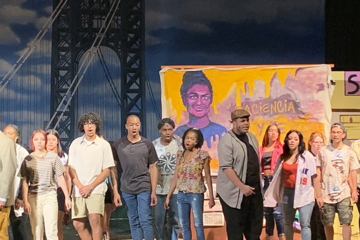 Several young people stand on a stage and sing in front of a backdrop of a bridge and a woman.