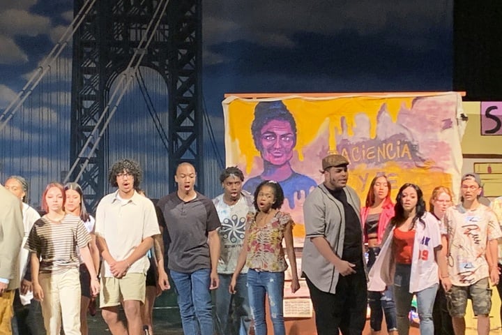 Several young people stand on a stage and sing in front of a backdrop of a bridge and a woman.