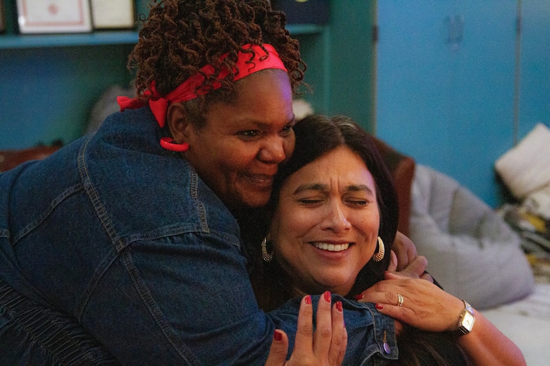 A woman with a read scarf and a jeans jacket hugs another woman with earrings.