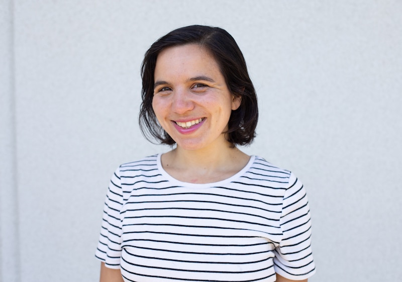 A photograph of a woman with short dark hair wearing a white shirt with black stripes posing for a portrait in front of a white wall.