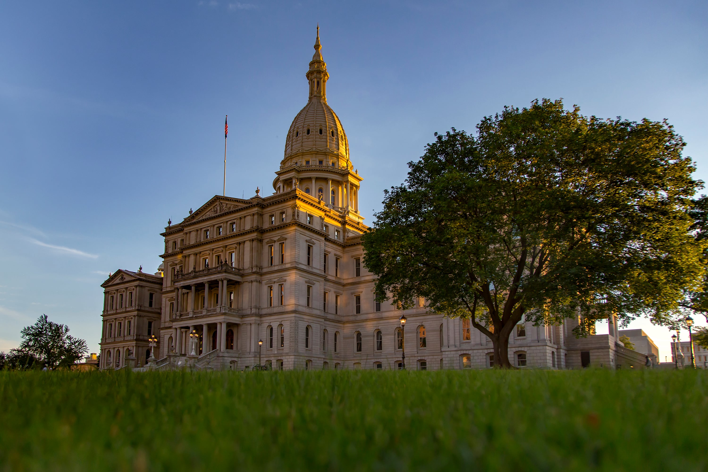 The sun is setting on the capitol building with a large green tree in the foreground and blue sky in the background.