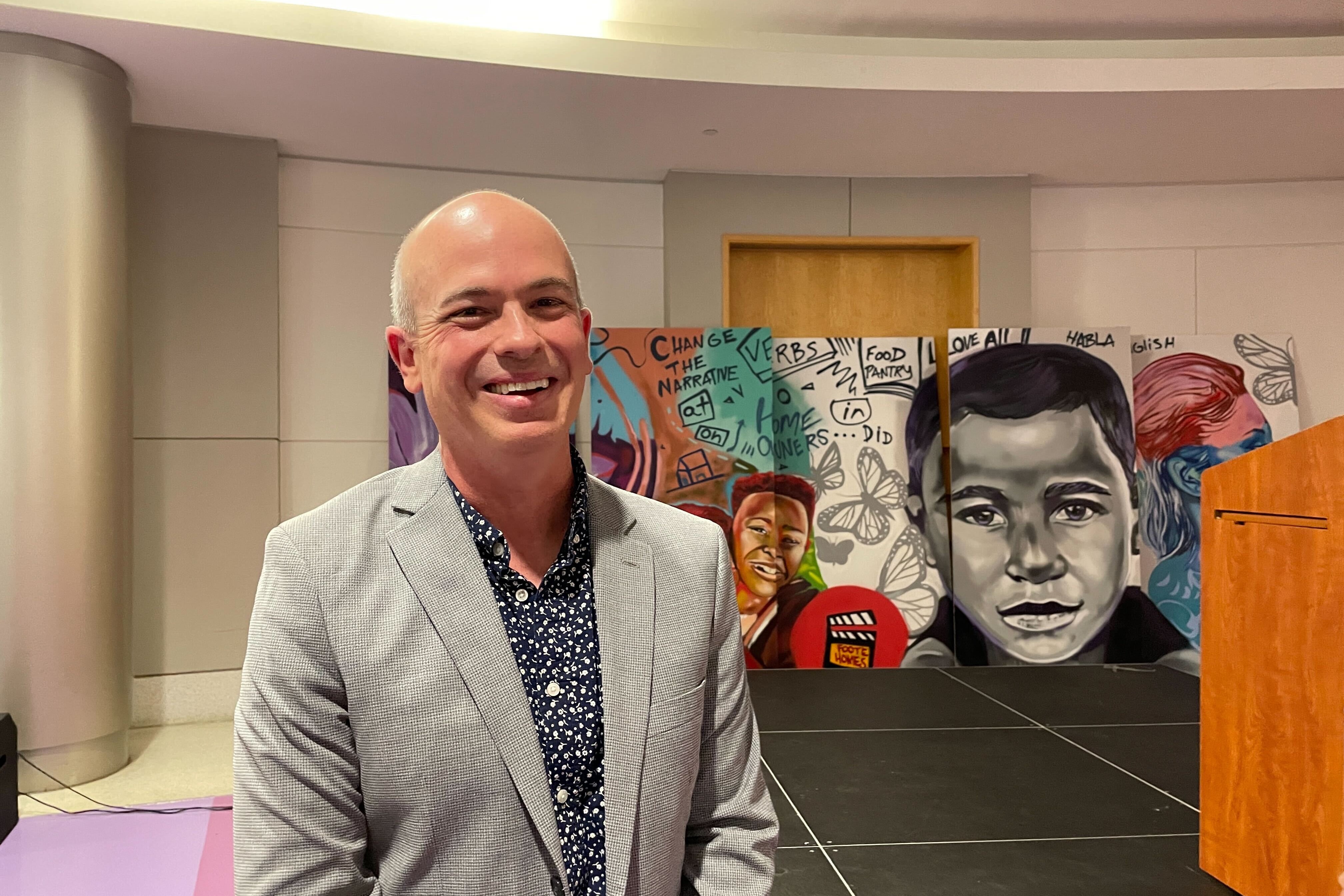 A man in a gray suit smiles for a portrait. In the background is artwork of a young boy.