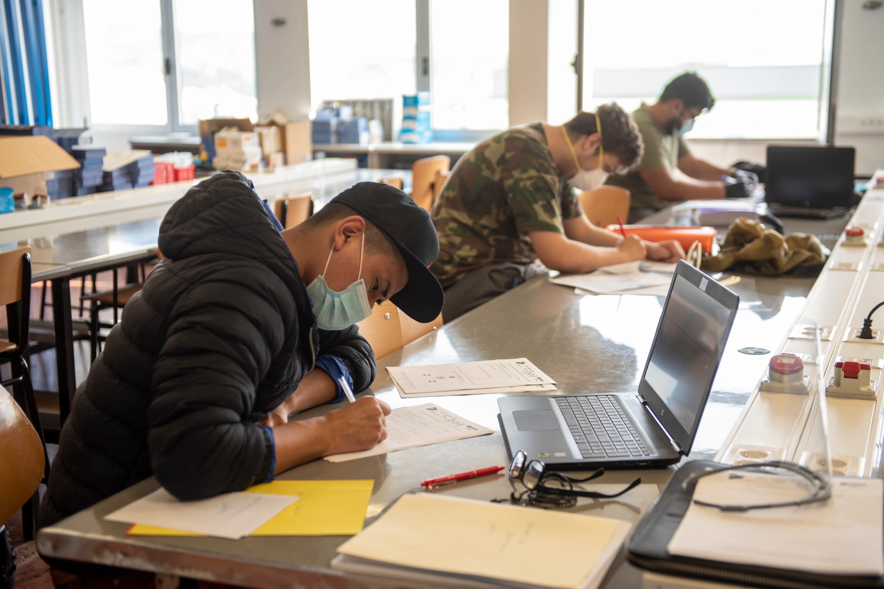 Three male students wearing masks sitting spaced out at a long table working on papers