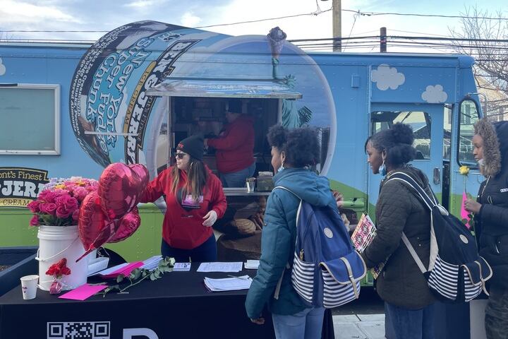 A truck with a Ben and Jerry’s pint is in the background while a woman in a red sweater gives a rose to a student.