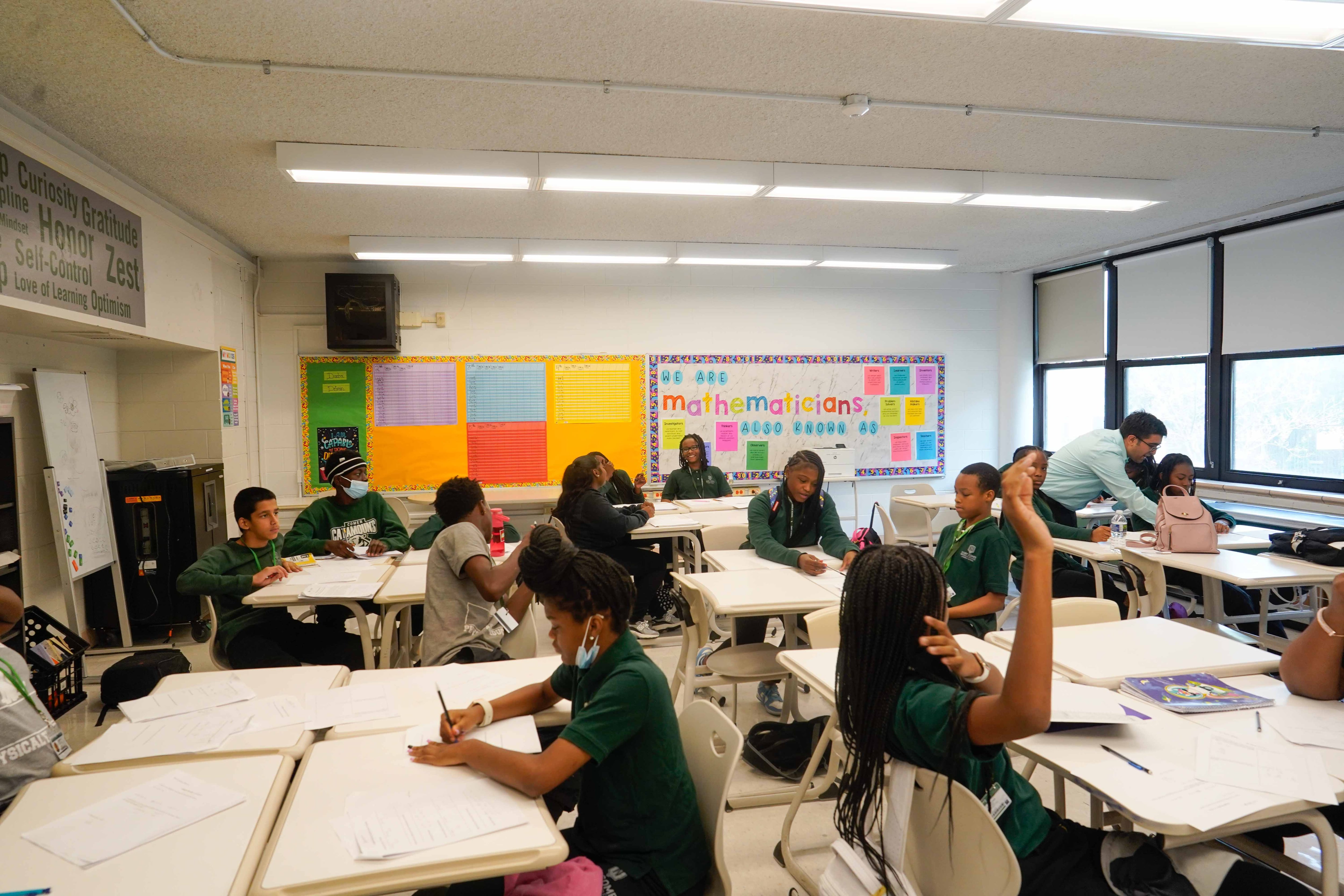 Students sit at groups of fours at tables. One student is raising a hand. A teacher is currently helping a student off to the side of a photo.