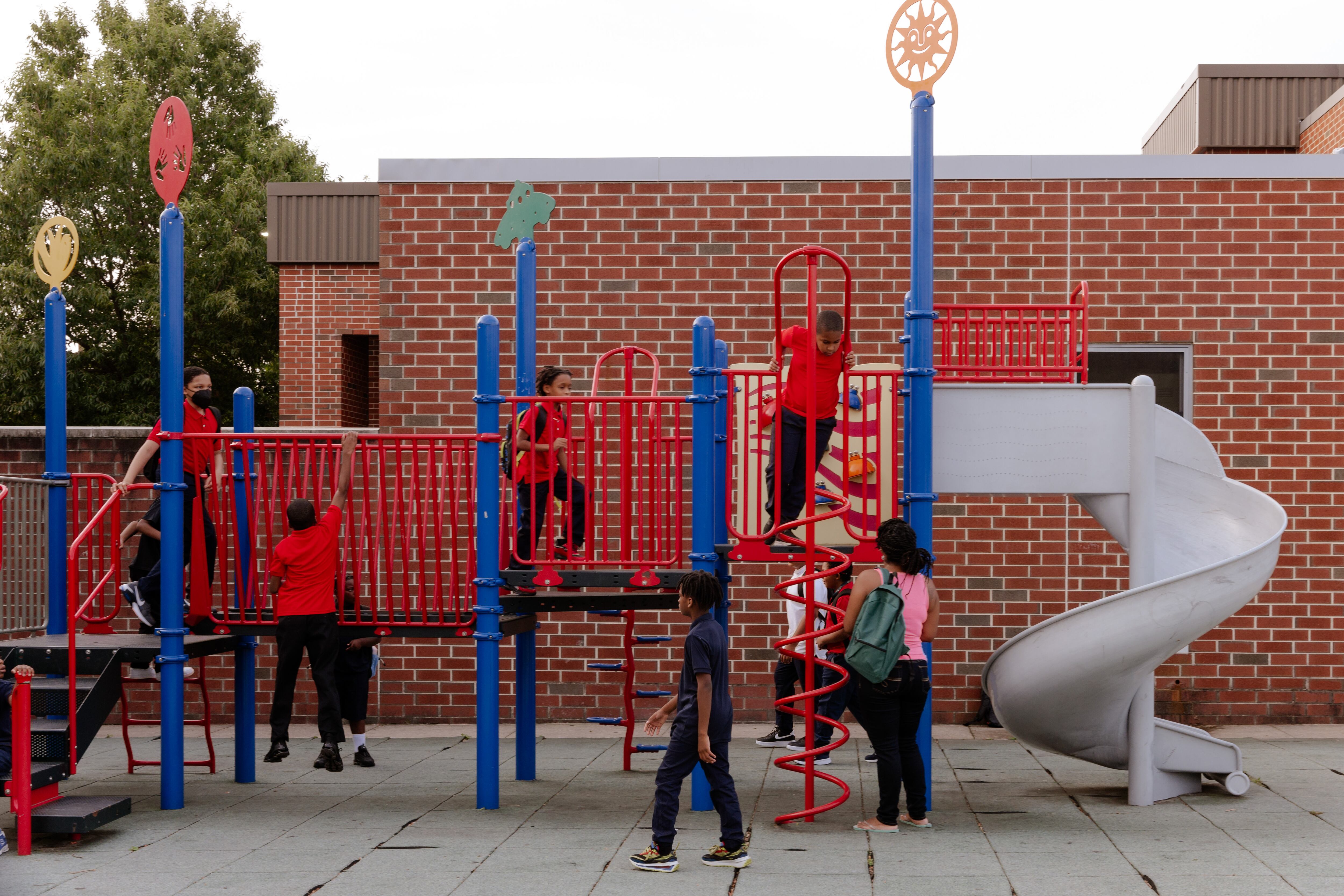 young students play on a red playground set with a school brick building in the background.