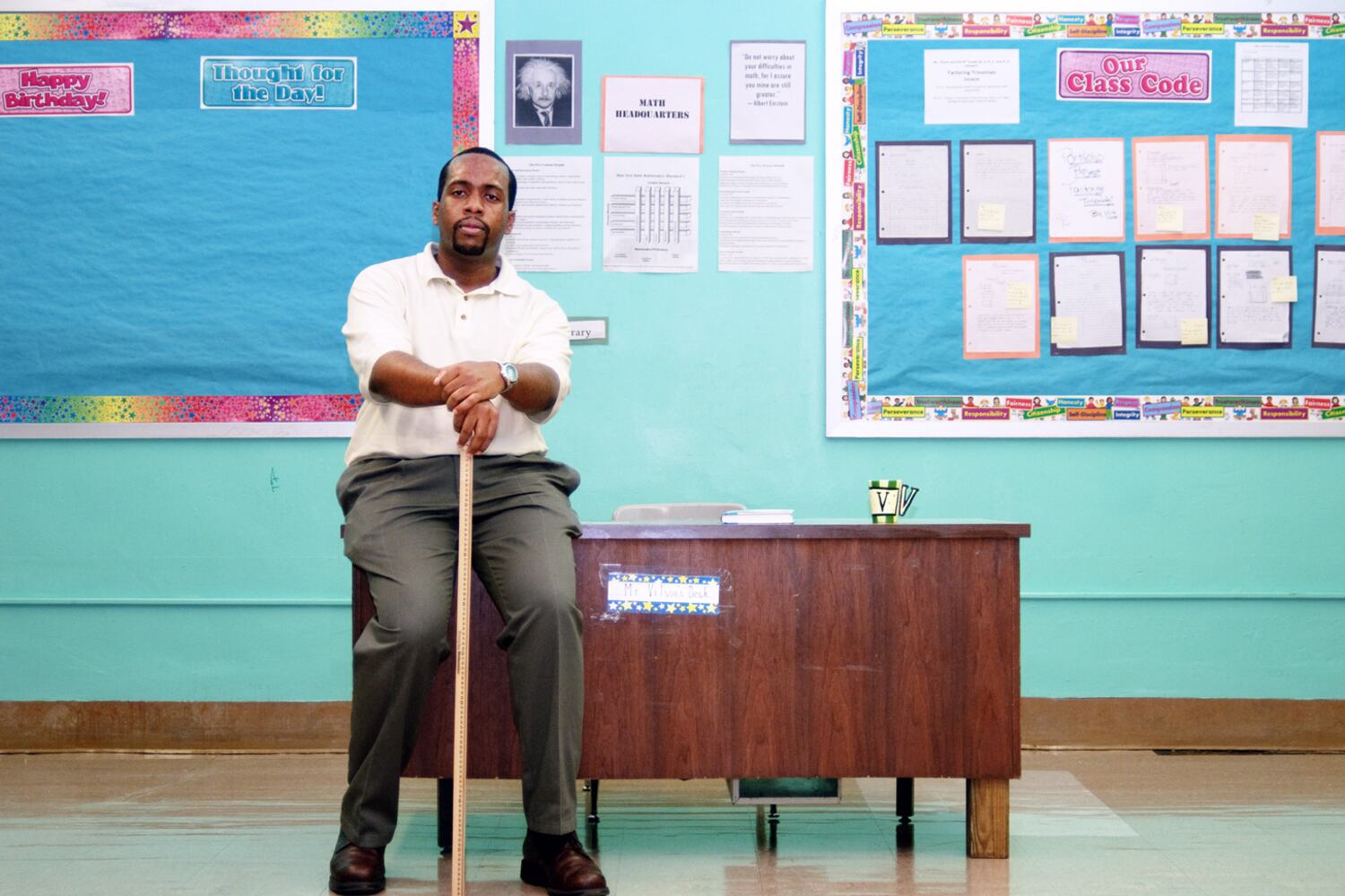 A man sits on a brown desk in a classroom. Blue bulletin boards are behind him.