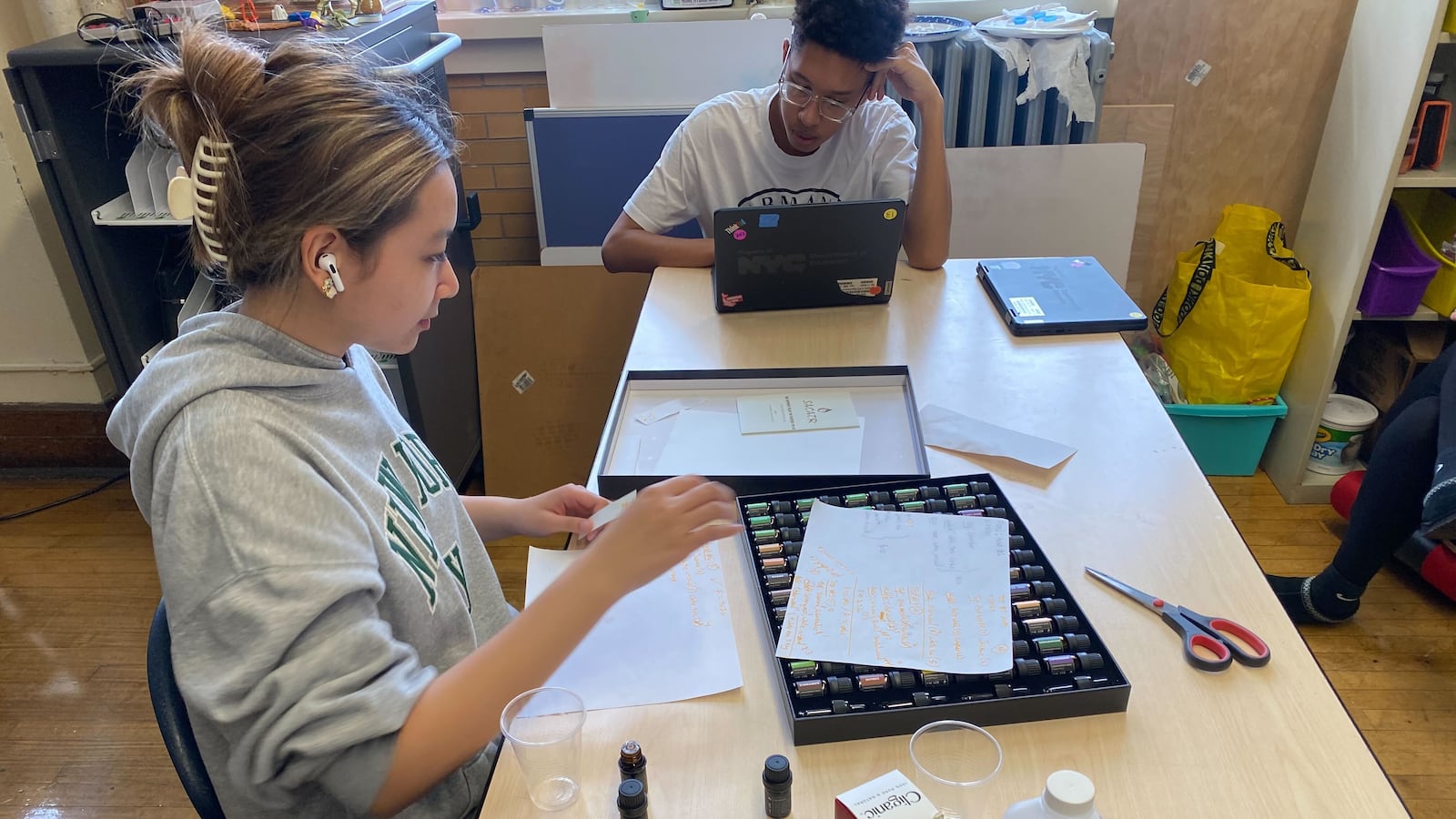 A girl sits at a table in a school over a case of essential oils. A pair of scissors, paper, and other materials are on the table.