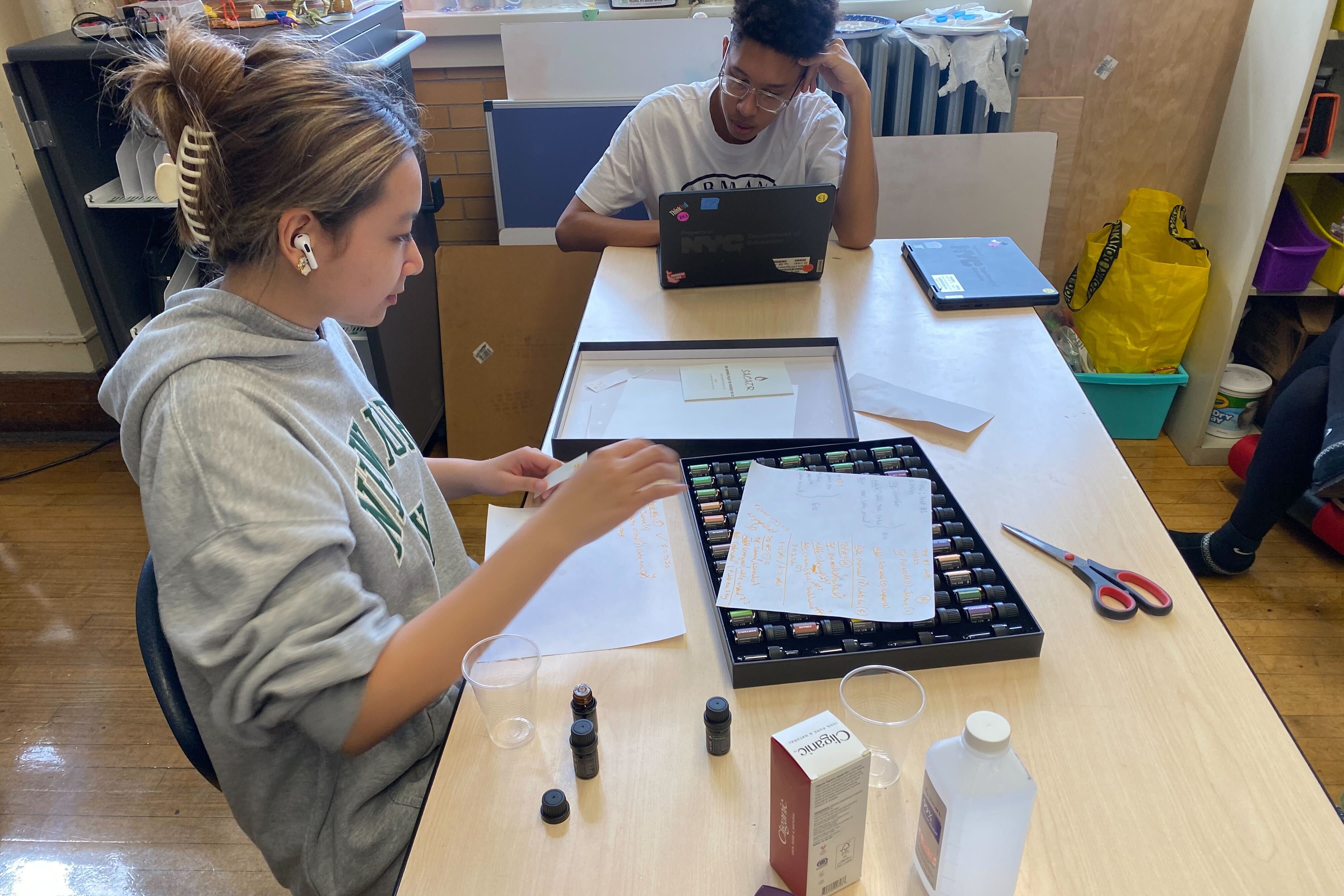 A girl sits at a table in a school over a case of essential oils. A pair of scissors, paper, and other materials are on the table.