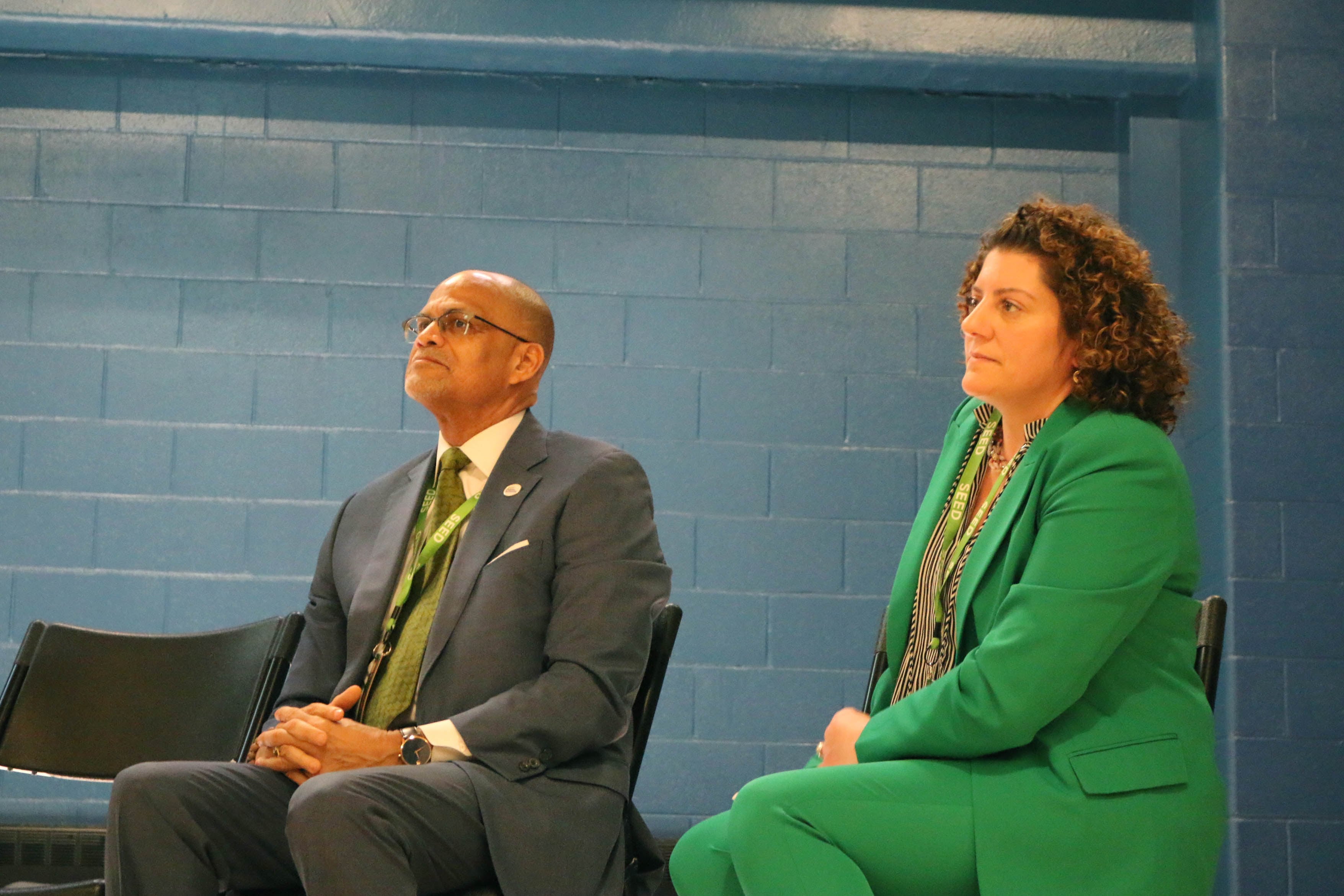 Two people wearing suits sit next to each other with a blue brick background.