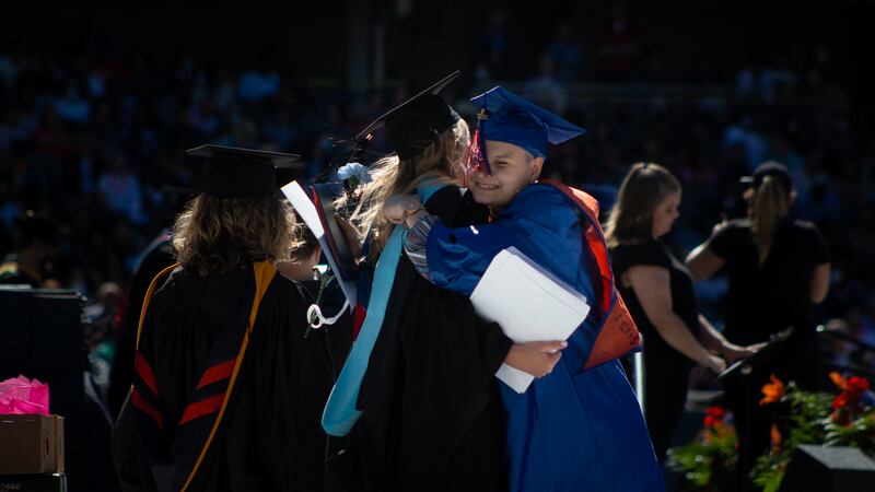 A young woman and educator embrace during a graduation ceremony, both wearing full graduation regalia.
