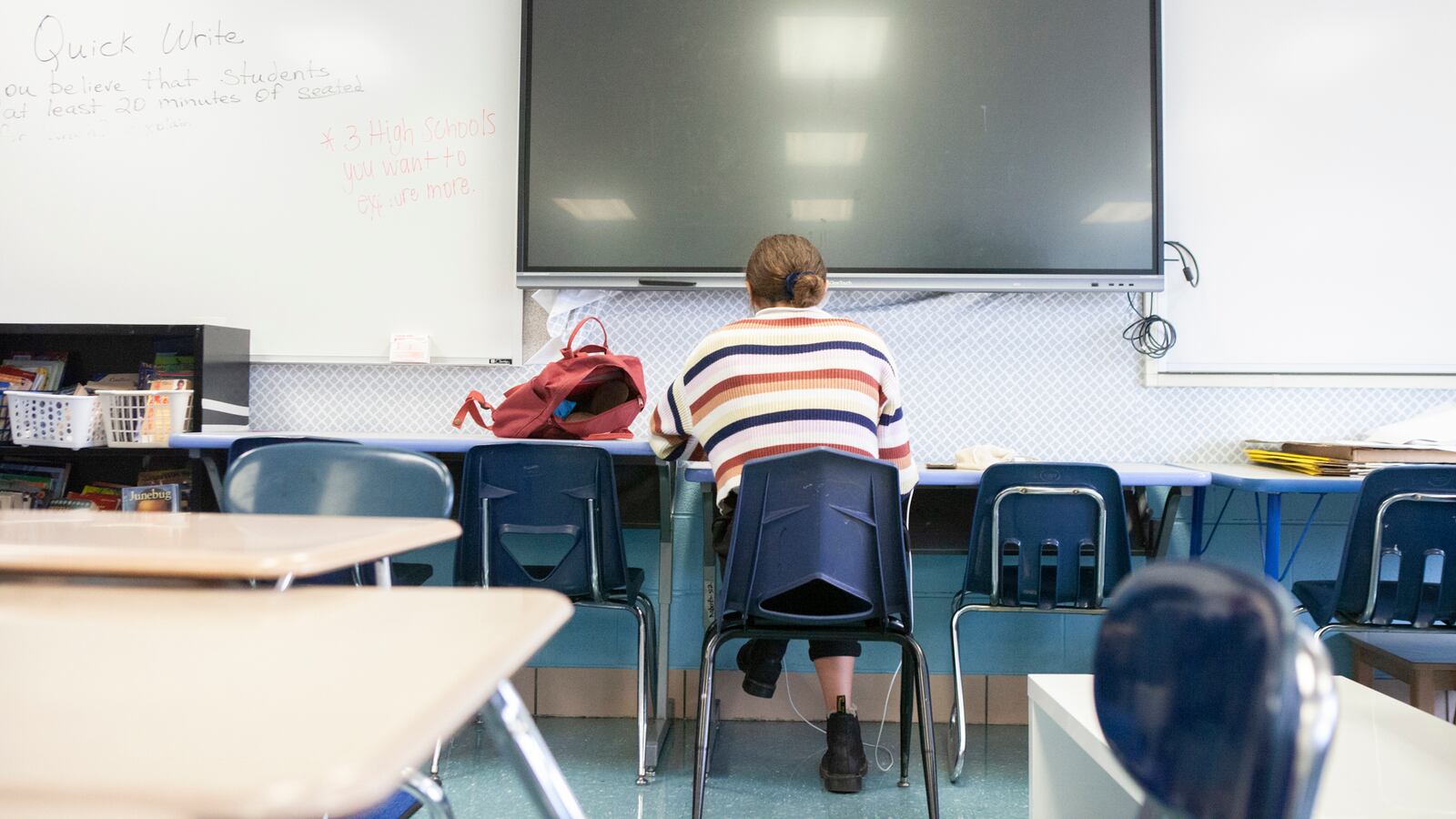 Back of a teacher sitting at a table in front of a large screen.
