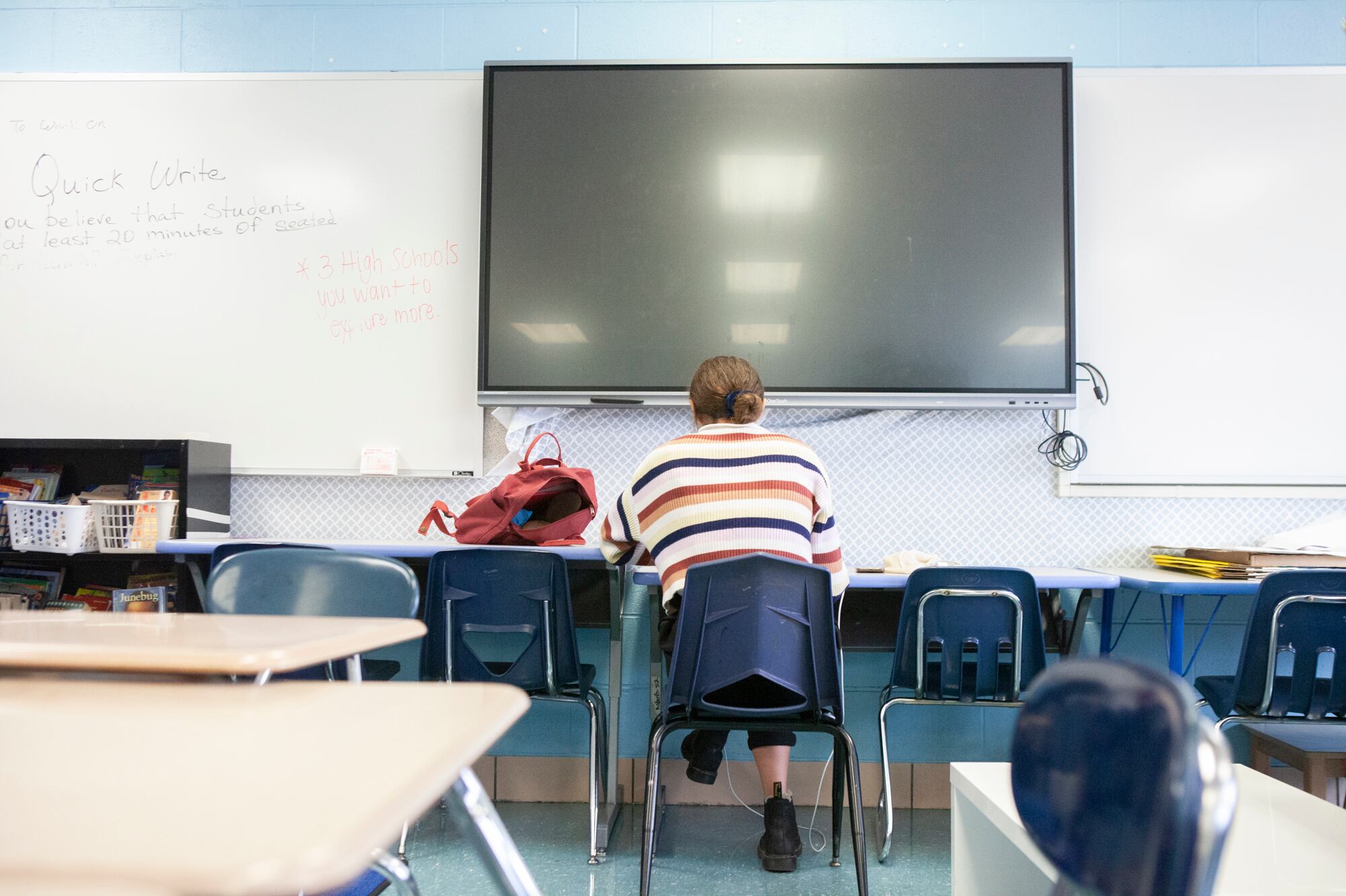 Back of a teacher sitting at a table in front of a large screen.